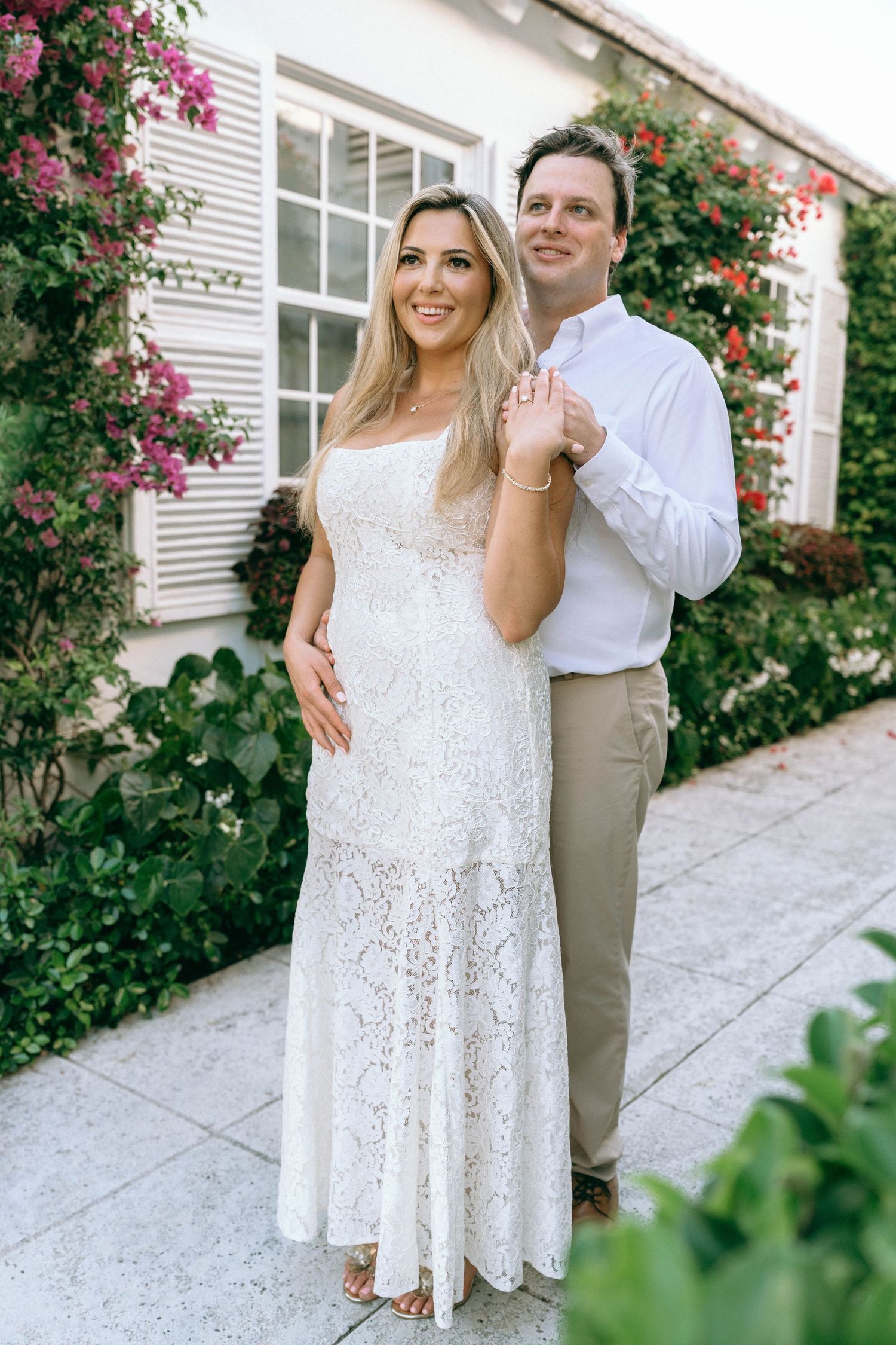 Couple posing outdoors in white lace dress and khaki pants near flowering bushes.