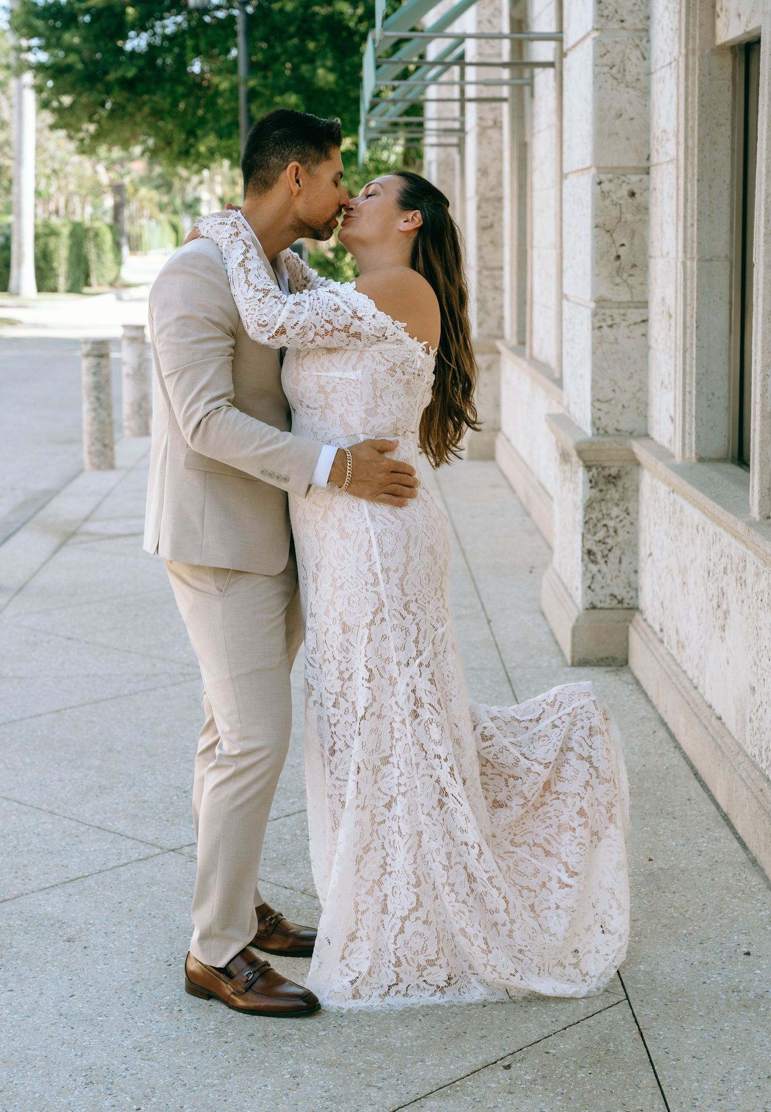 Bride in lace gown and groom in tan suit share a kiss on a sunlit sidewalk outside a stone building.