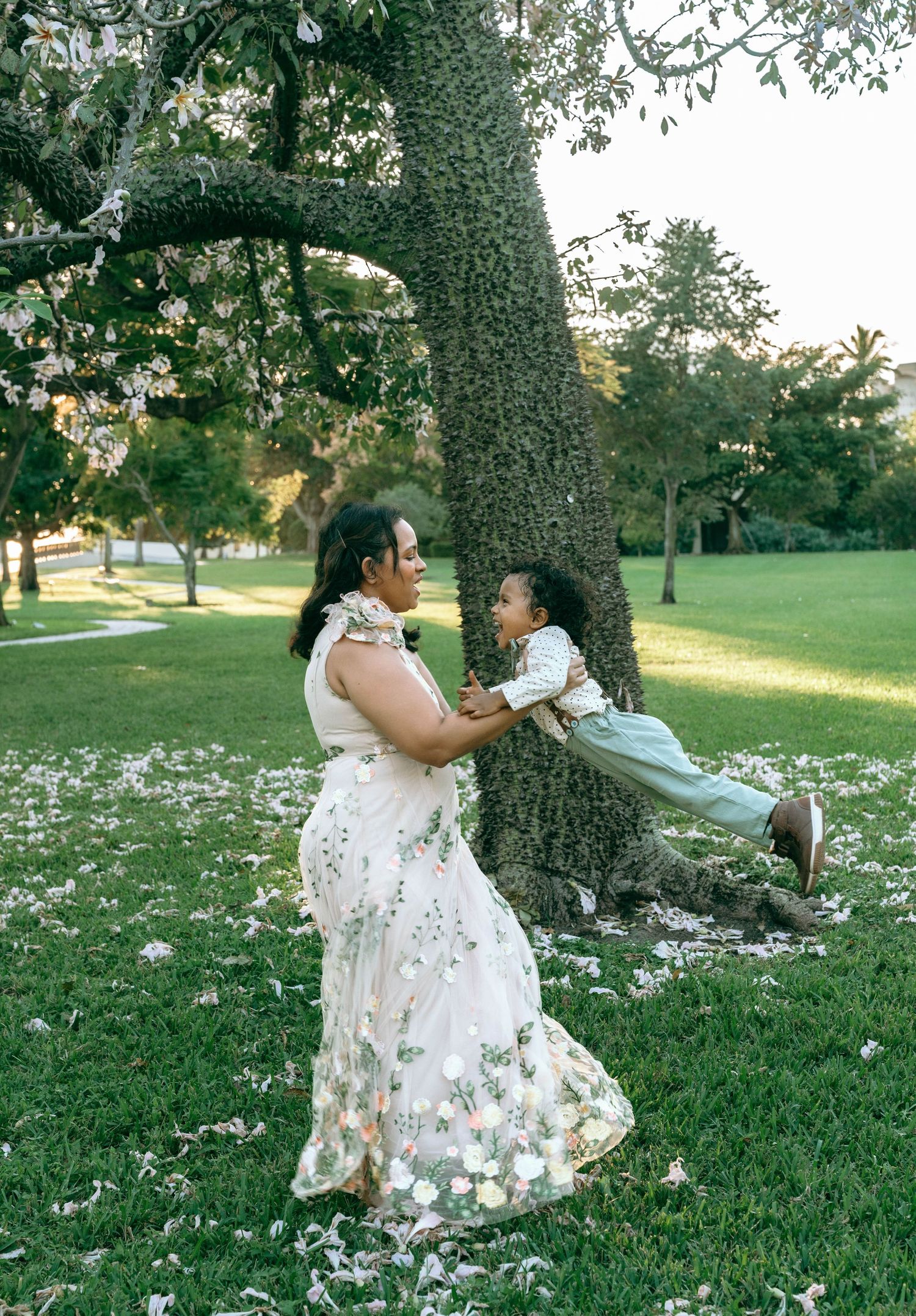 A person in a floral white dress sits under a large tree in a grassy field scattered with white flower petals.