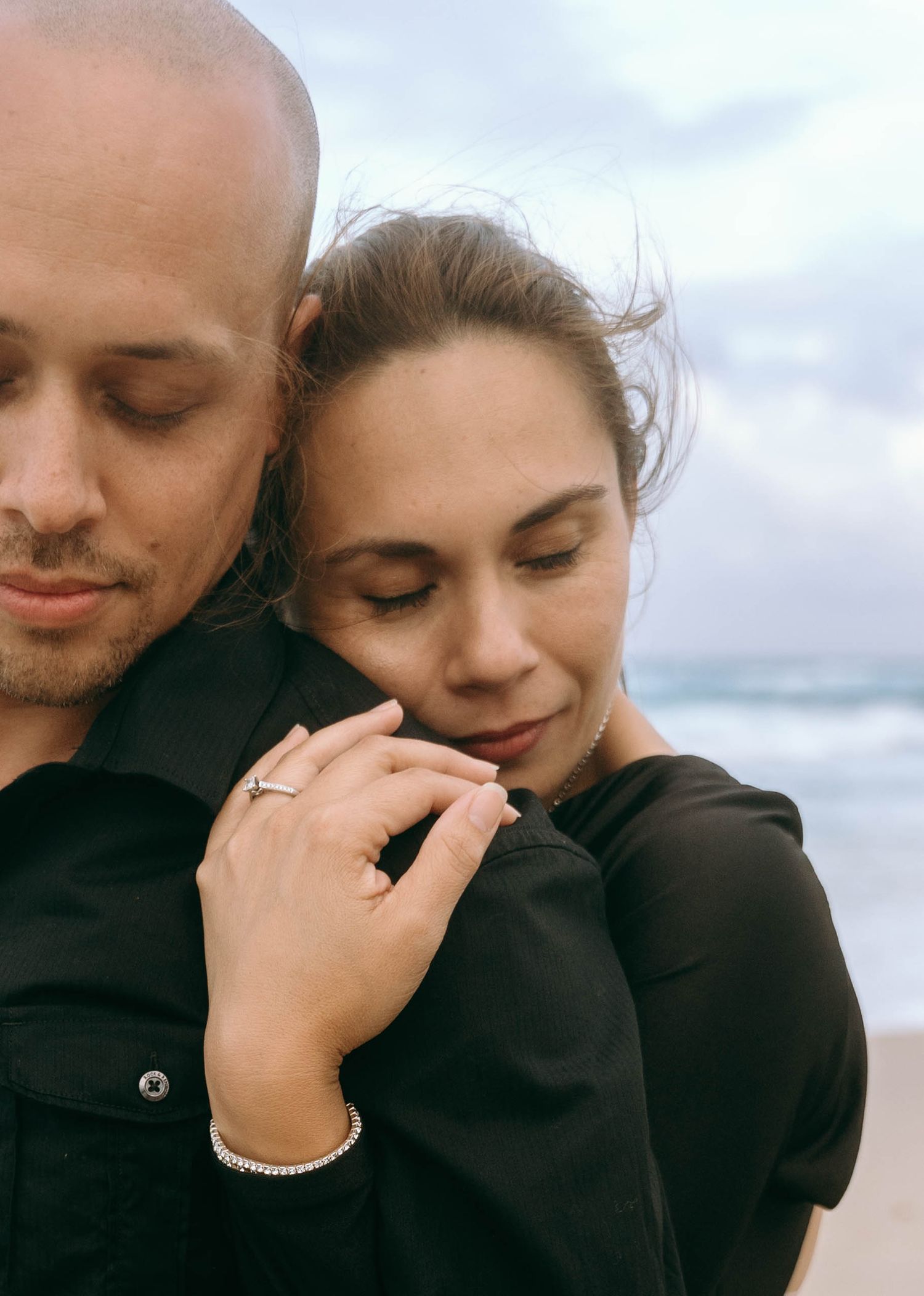 Close-up of couple in dark clothing sharing intimate moment on cloudy beach.