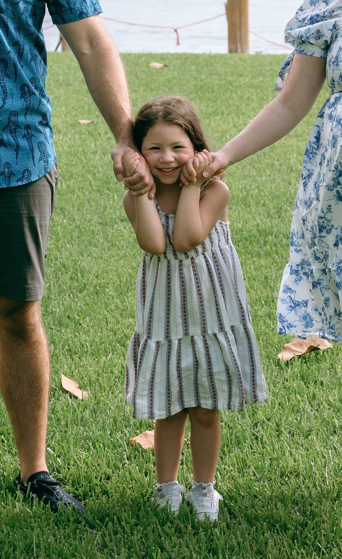 A smiling young girl in a striped dress holds hands with her parents while standing on green grass outdoors.