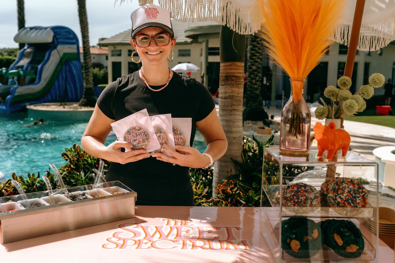 Woman smiling at a candy dessert table by a pool, holding packaged treats at an outdoor party event.