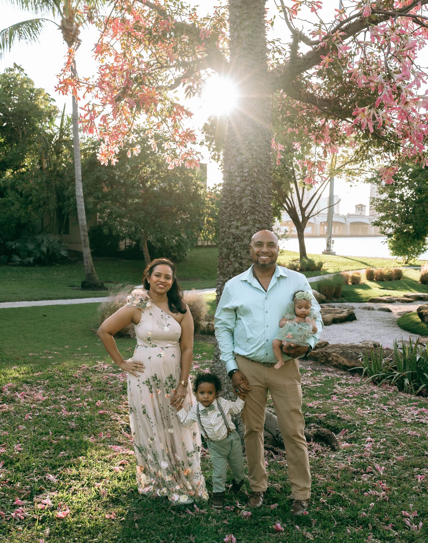 Family of four posing outdoors under blooming trees with sunlight filtering through, in a lush green park setting at Bradley Park in Palm B.
