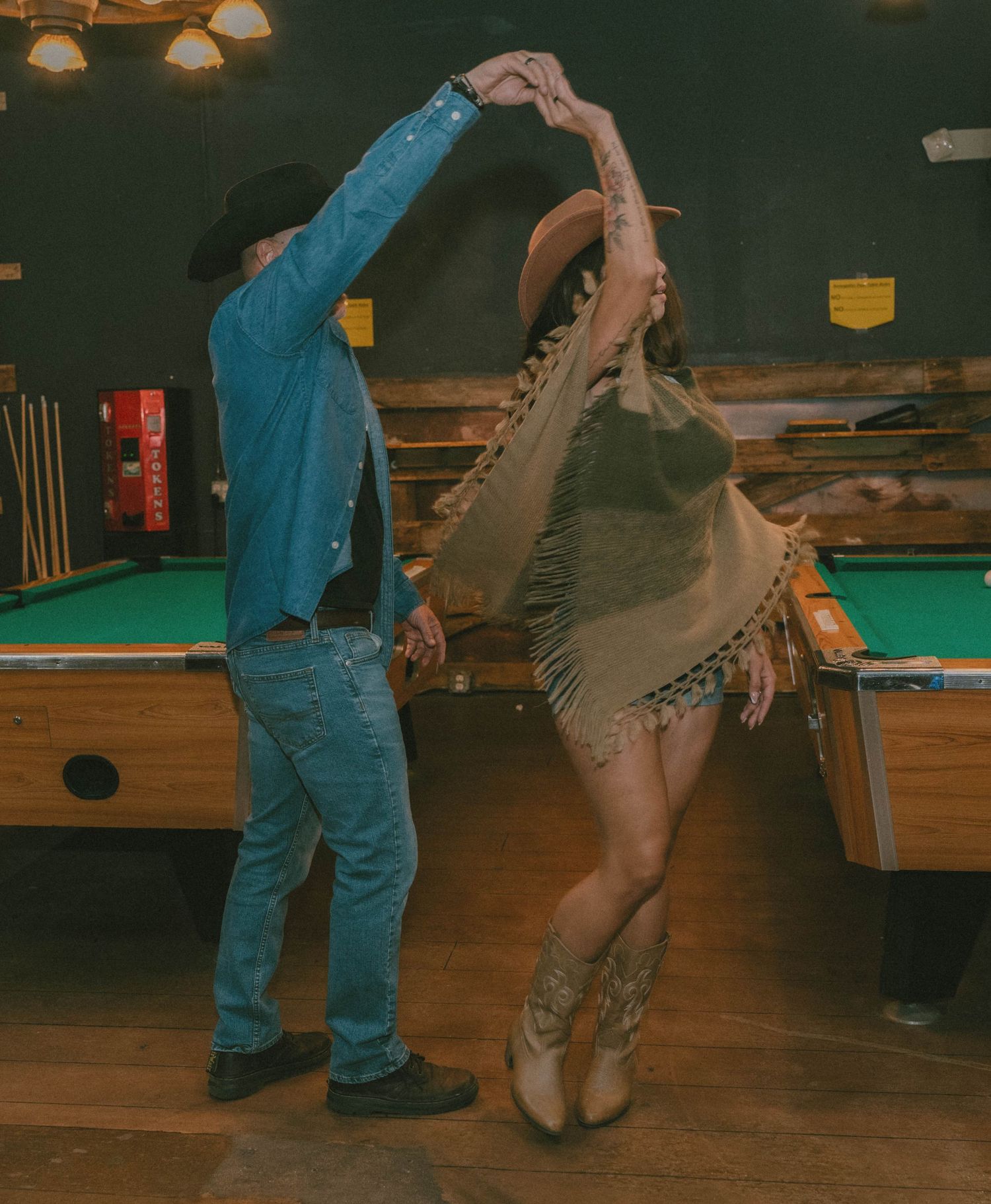 Couple line dancing in cowboy attire near pool tables in a rustic bar setting.
