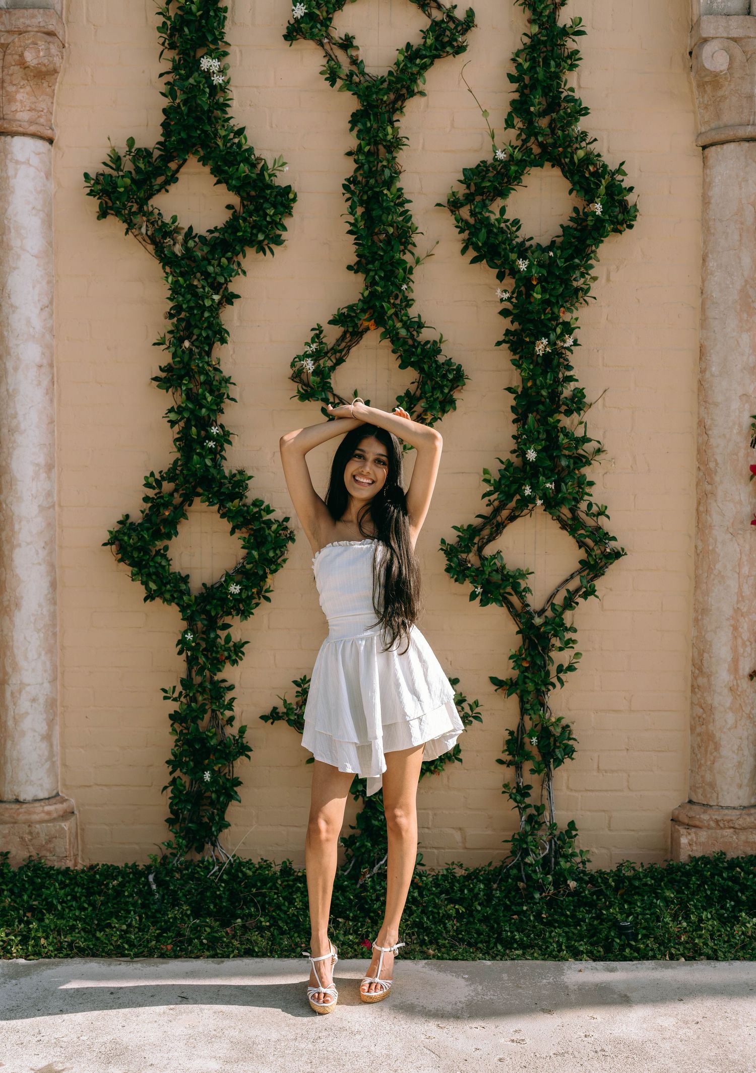 Young woman in white strapless mini dress posing with arms raised before an ivy-covered beige wall on Worth Avenue Palm beach.