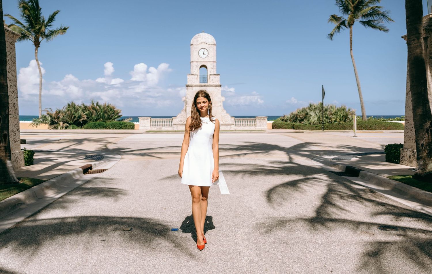 Woman in white dress standing on palm-lined path with ocean view and clock tower landmark in background on Worth Avenue Palm beach.