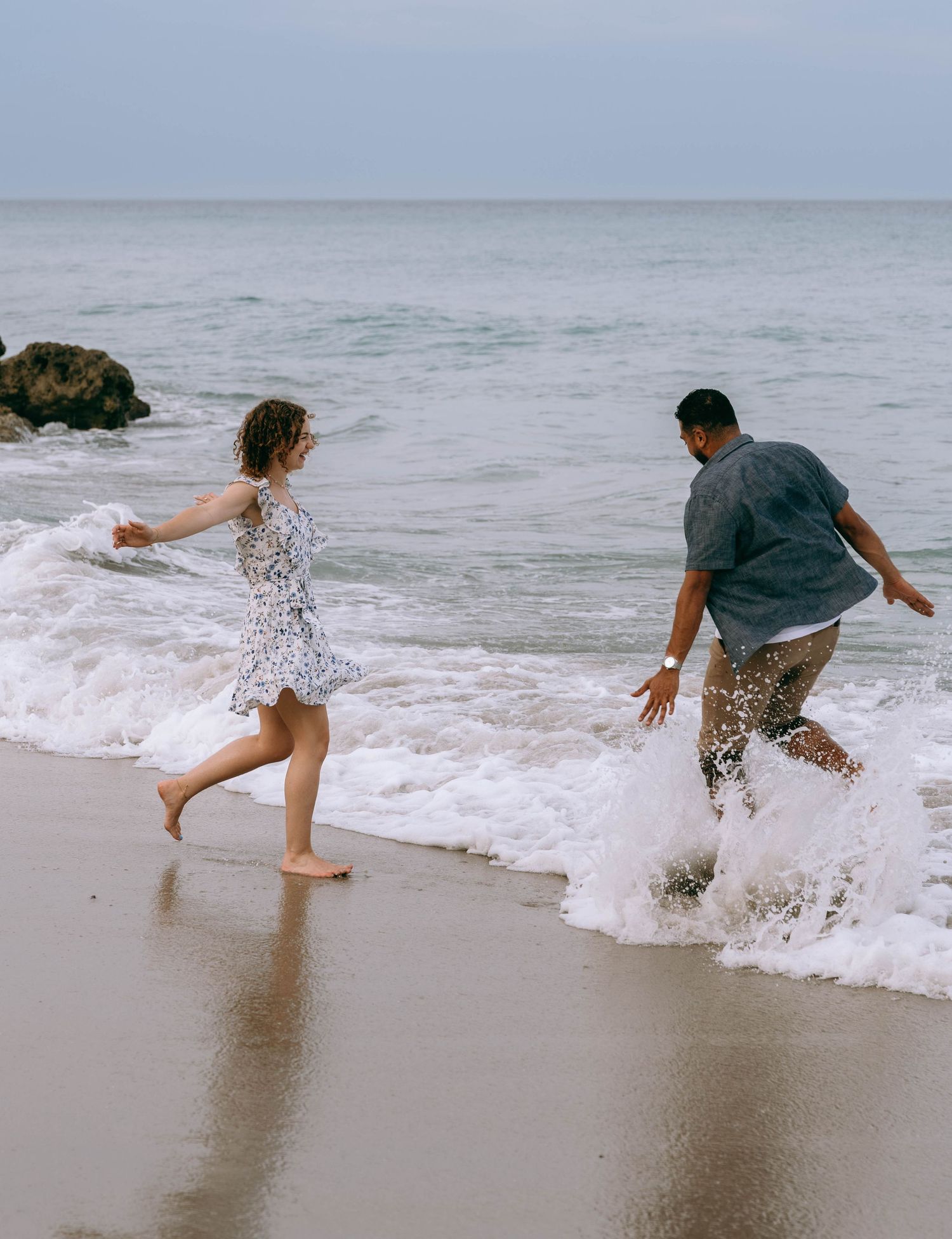 A couple playfully splashing in ocean waves on a sandy beach, woman in floral dress and man in casual shirt. Worth Avenue, Palm Beach, FL