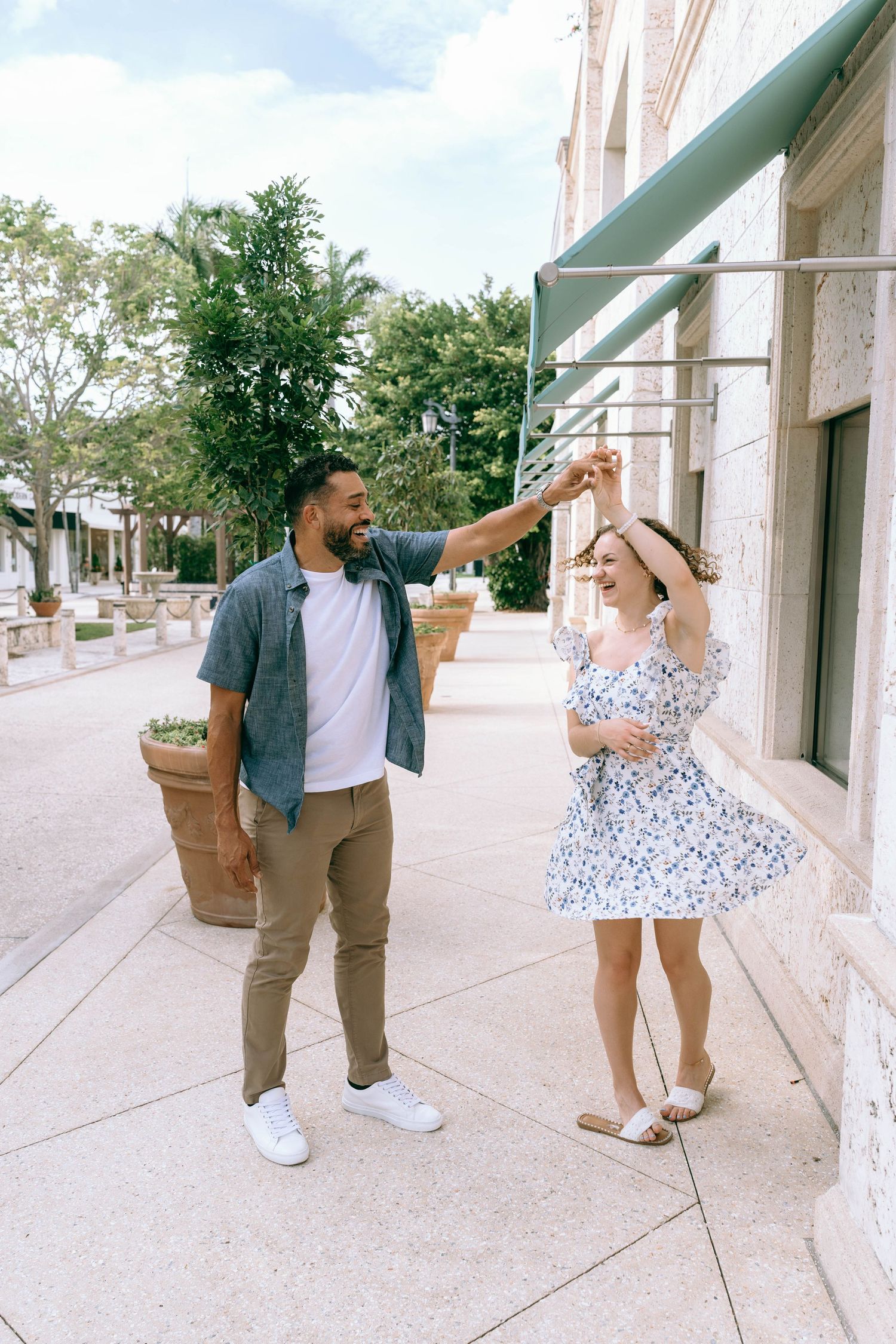 A joyful interracial couple dancing on a sunny outdoor shopping promenade, man spinning woman in floral dress. Engagement photos Worth AVe