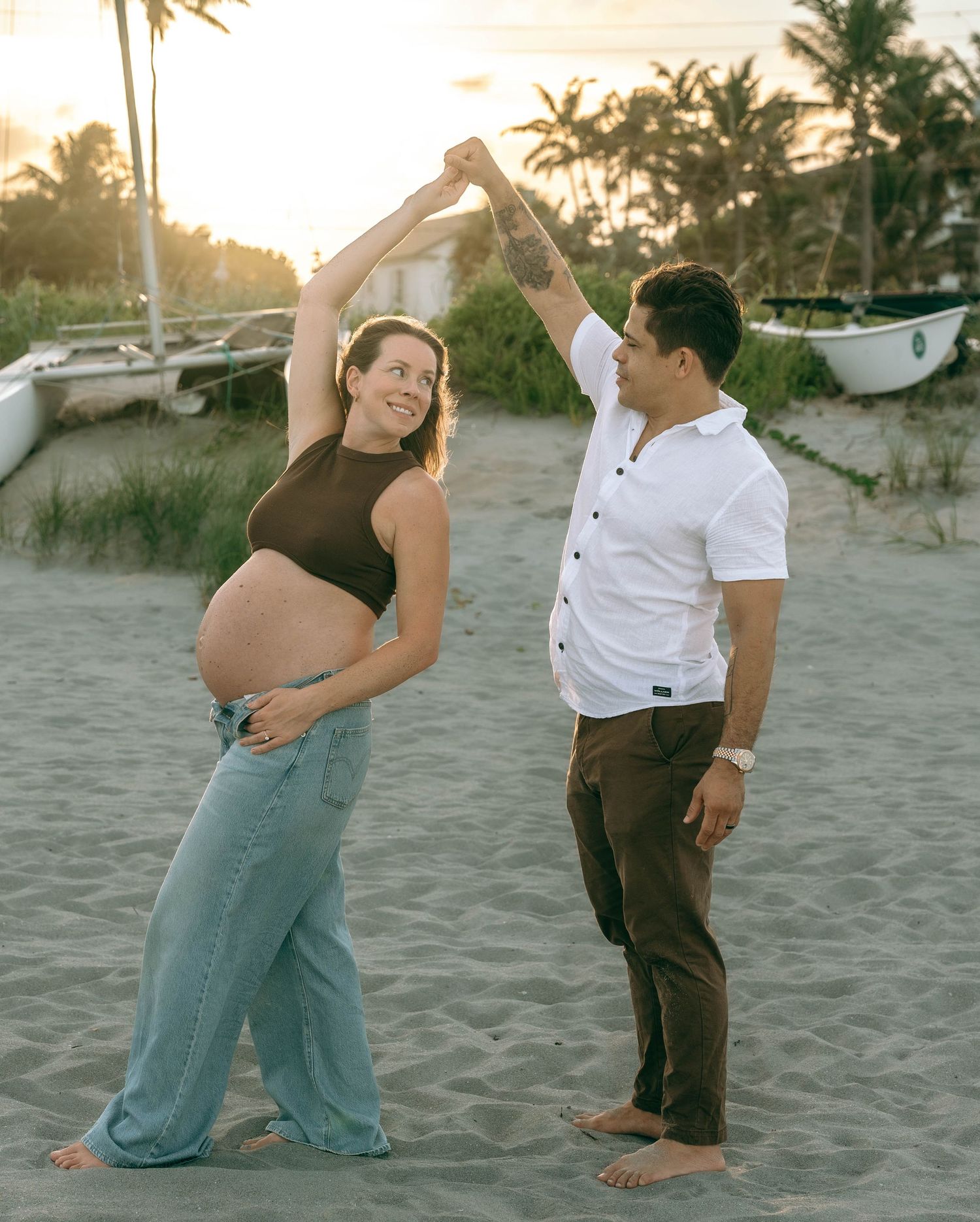 Pregnant couple dancing on beach at sunset, woman showing baby bump in crop top and jeans, man in white shirt.