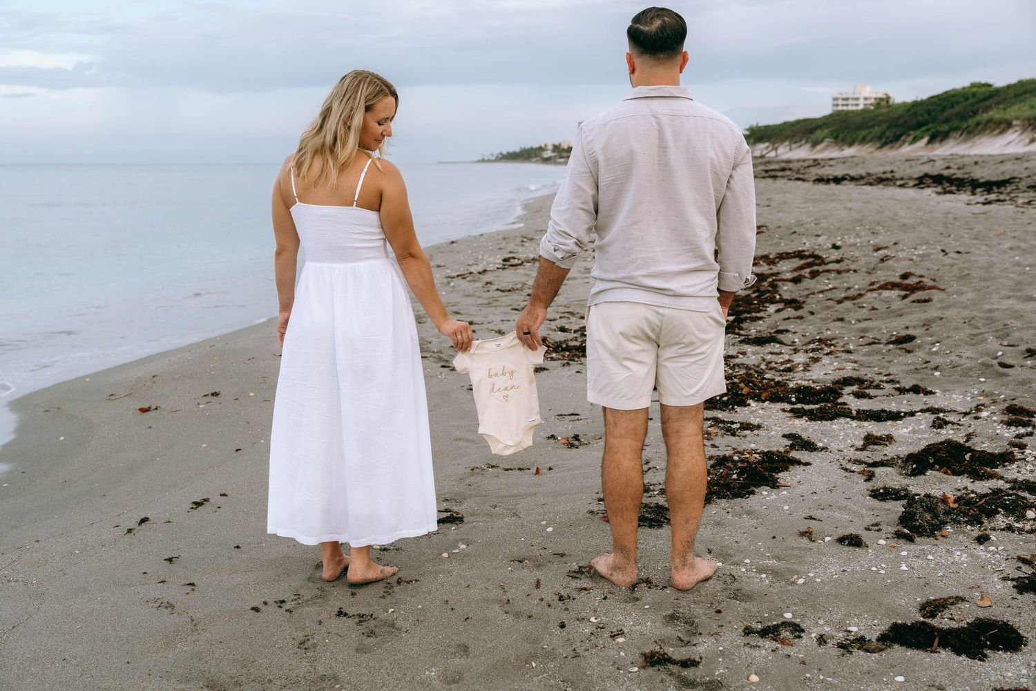 Couple in white outfits holding baby shoes while walking on a sandy beach at dusk.