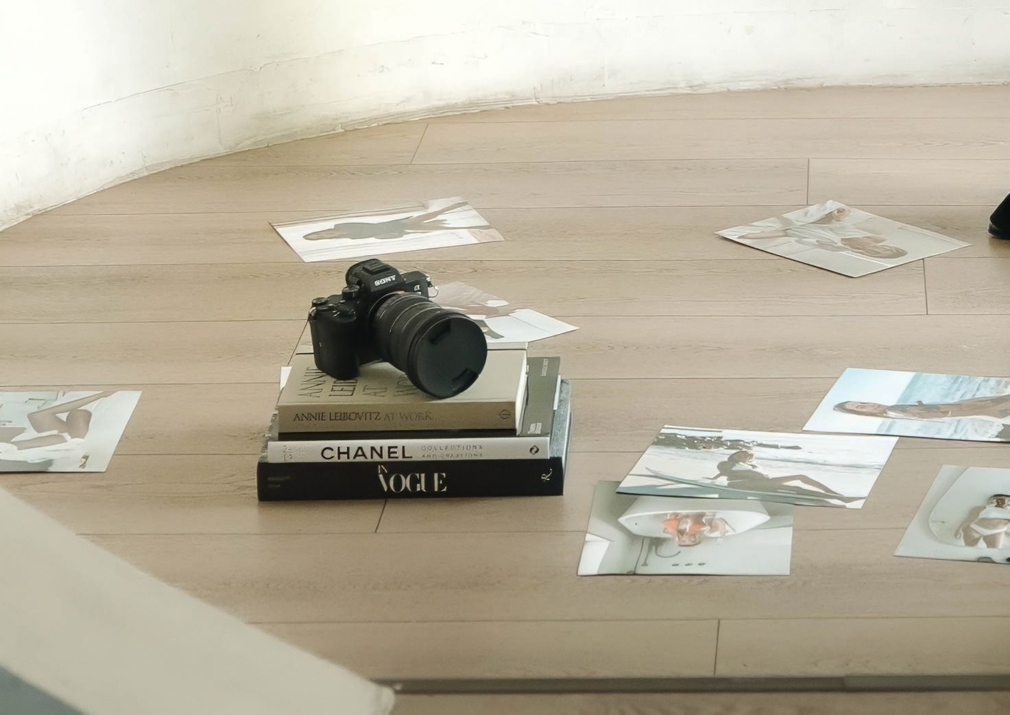A camera sits atop fashion books with scattered photographs on wooden floor.