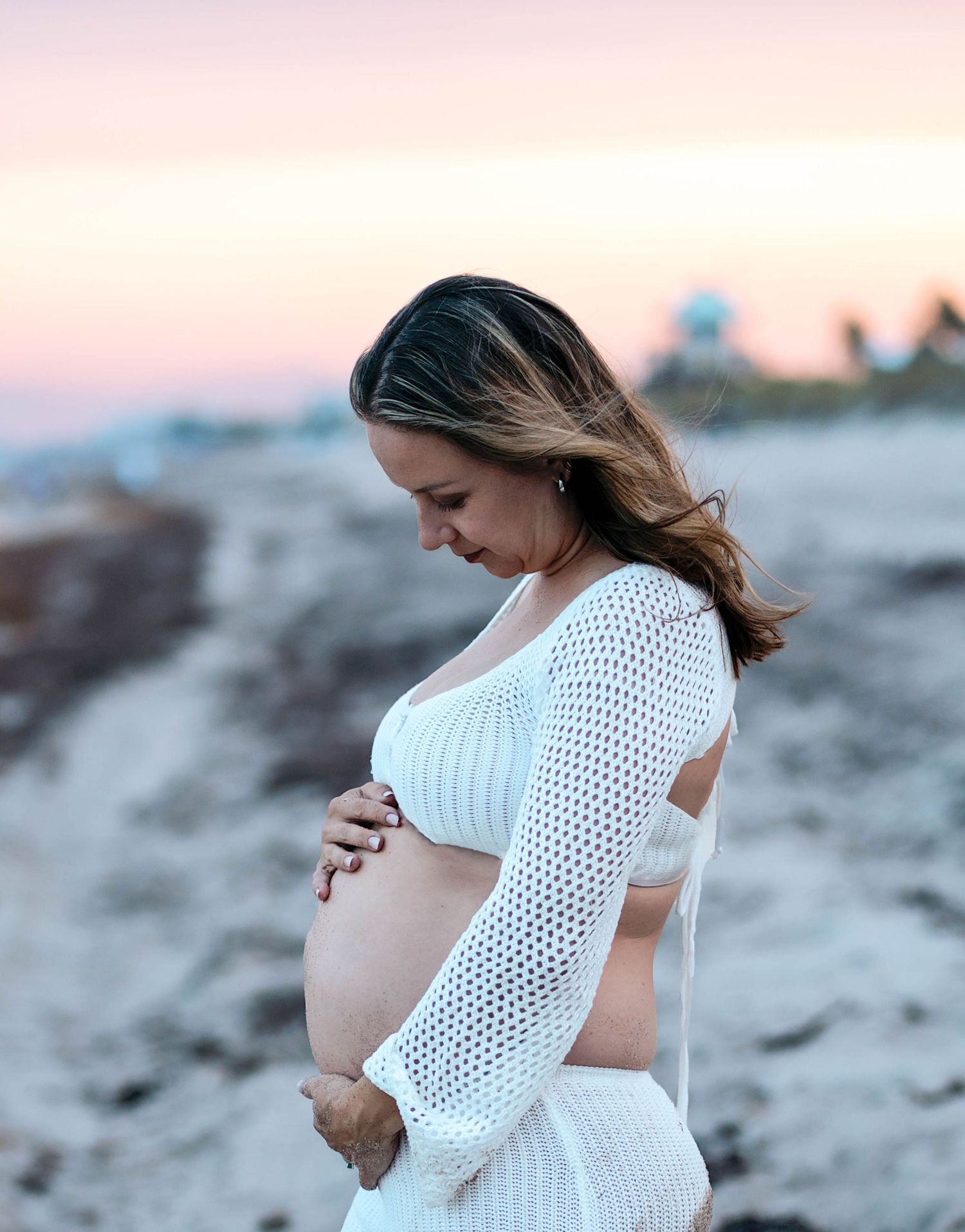 A woman in a white crochet dress poses on the beach during sunset.
