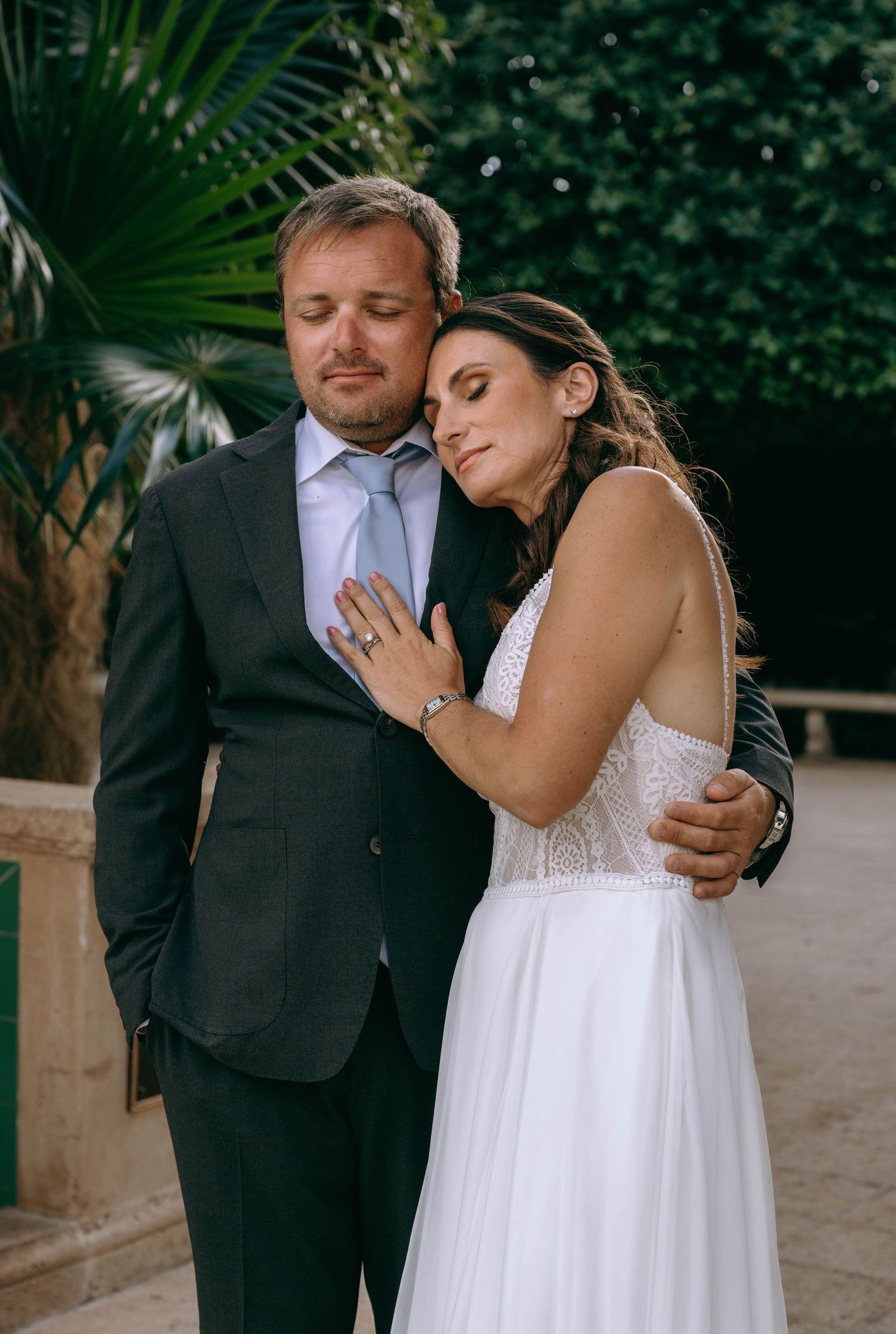 Elegant bride in white gown rests head on groom's chest in dark suit, posing outdoors near tropical greenery.