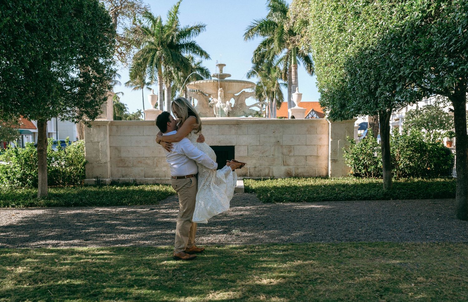 Groom lifting bride in white dress outdoors near a fountain surrounded by lush tropical palm trees.