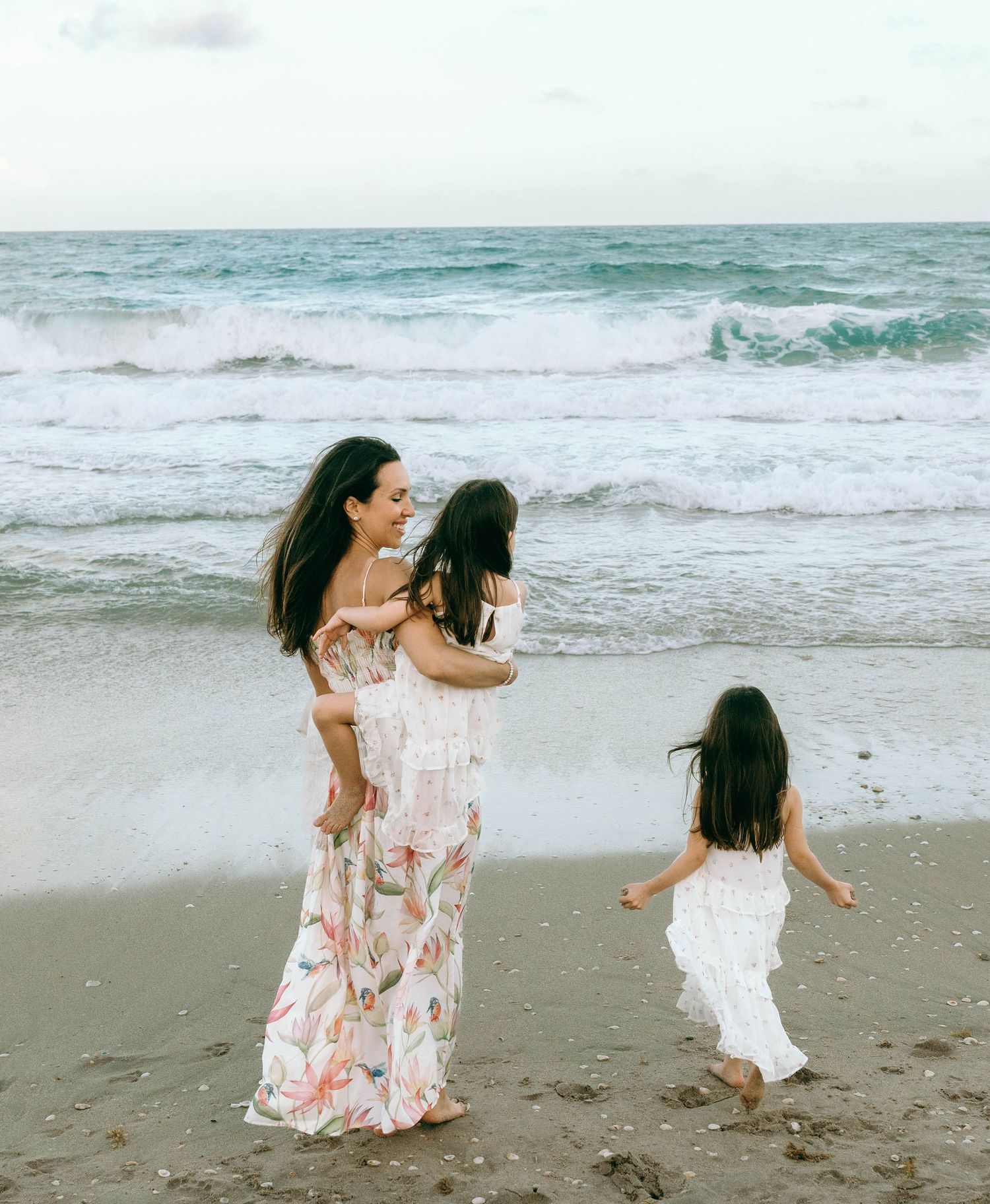 Mother in floral maxi dress holds daughter while another child runs toward ocean waves on sandy beach. Worth Avenue, Palm Beach, FL