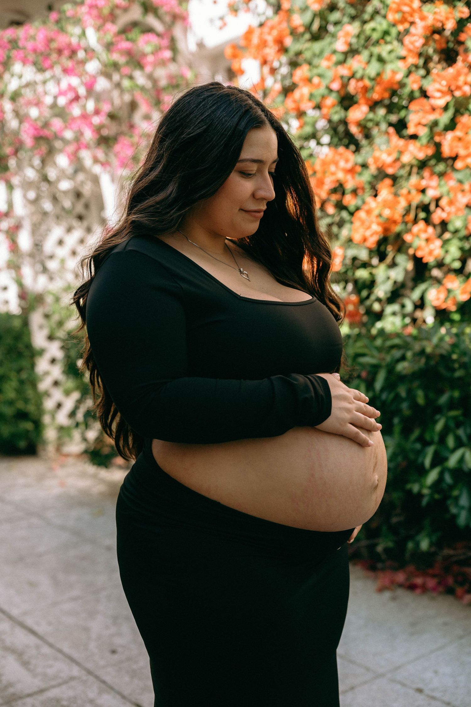 Pregnant woman in black dress cradling baby bump outdoors with colorful bougainvillea flowers in background. Worth Avenue, Palm Beach, FL