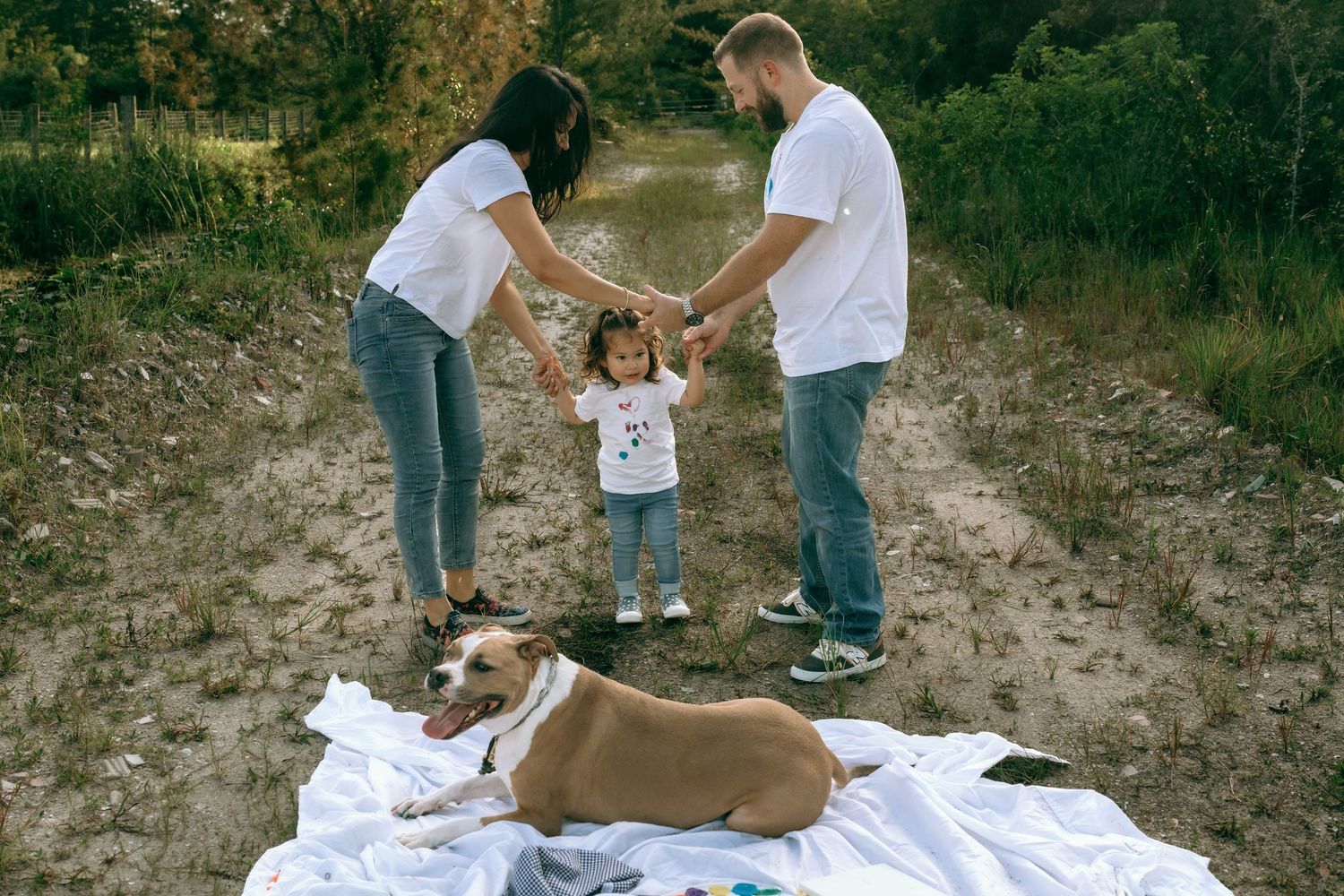 Family with young child and dog enjoying outdoor picnic on white blanket in nature trail.
