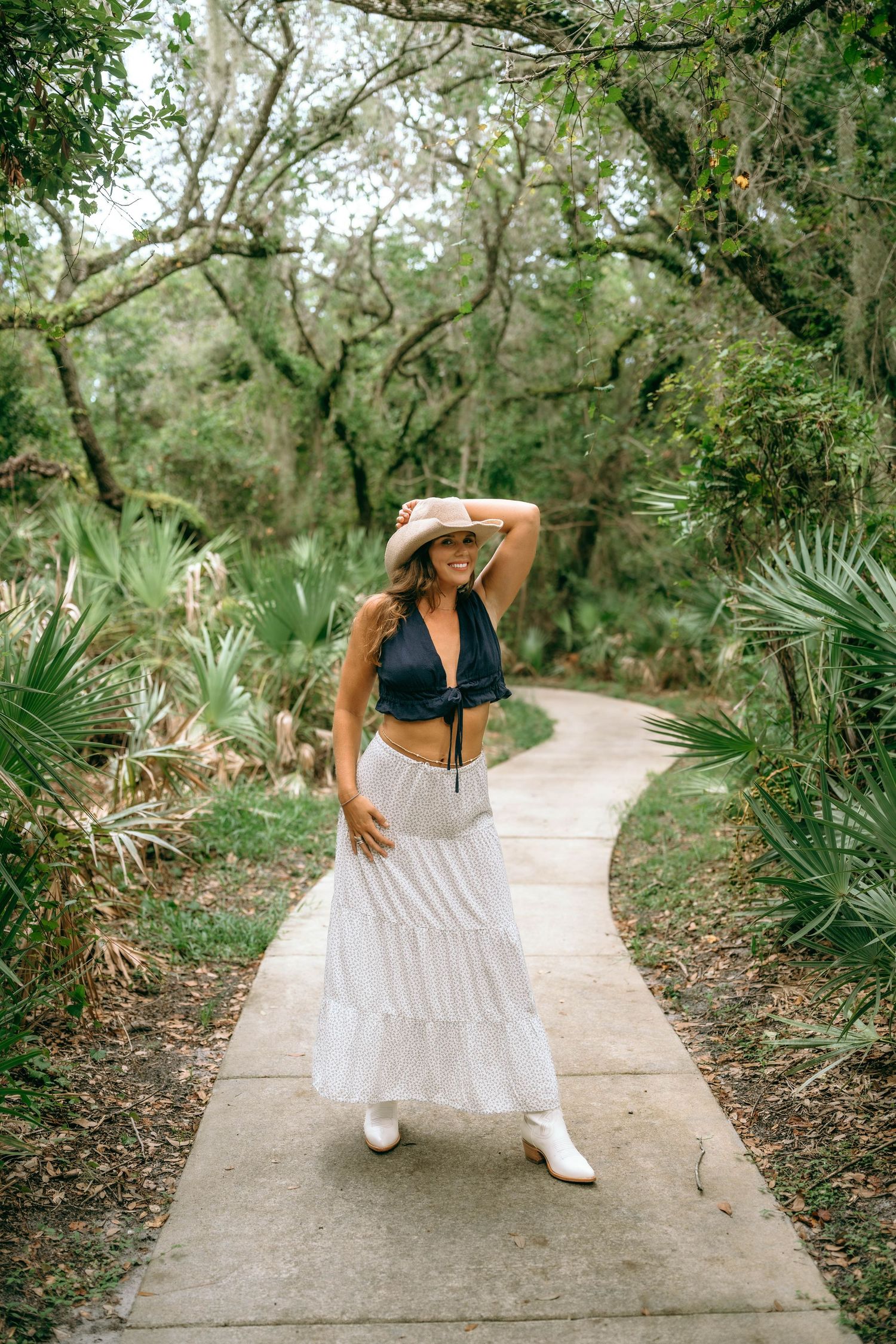 Woman in white maxi skirt, crop top, and cowboy hat posing on a garden path surrounded by lush greenery.