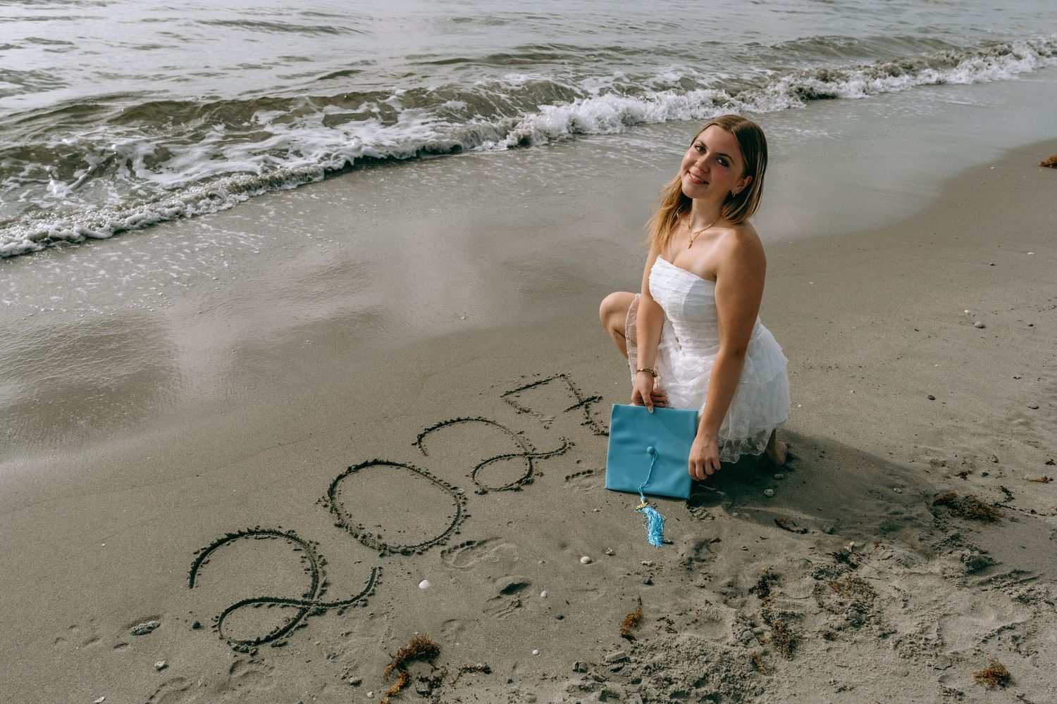 Woman in white dress sitting on beach sand next to '2023' written in sand, holding blue graduation cap.