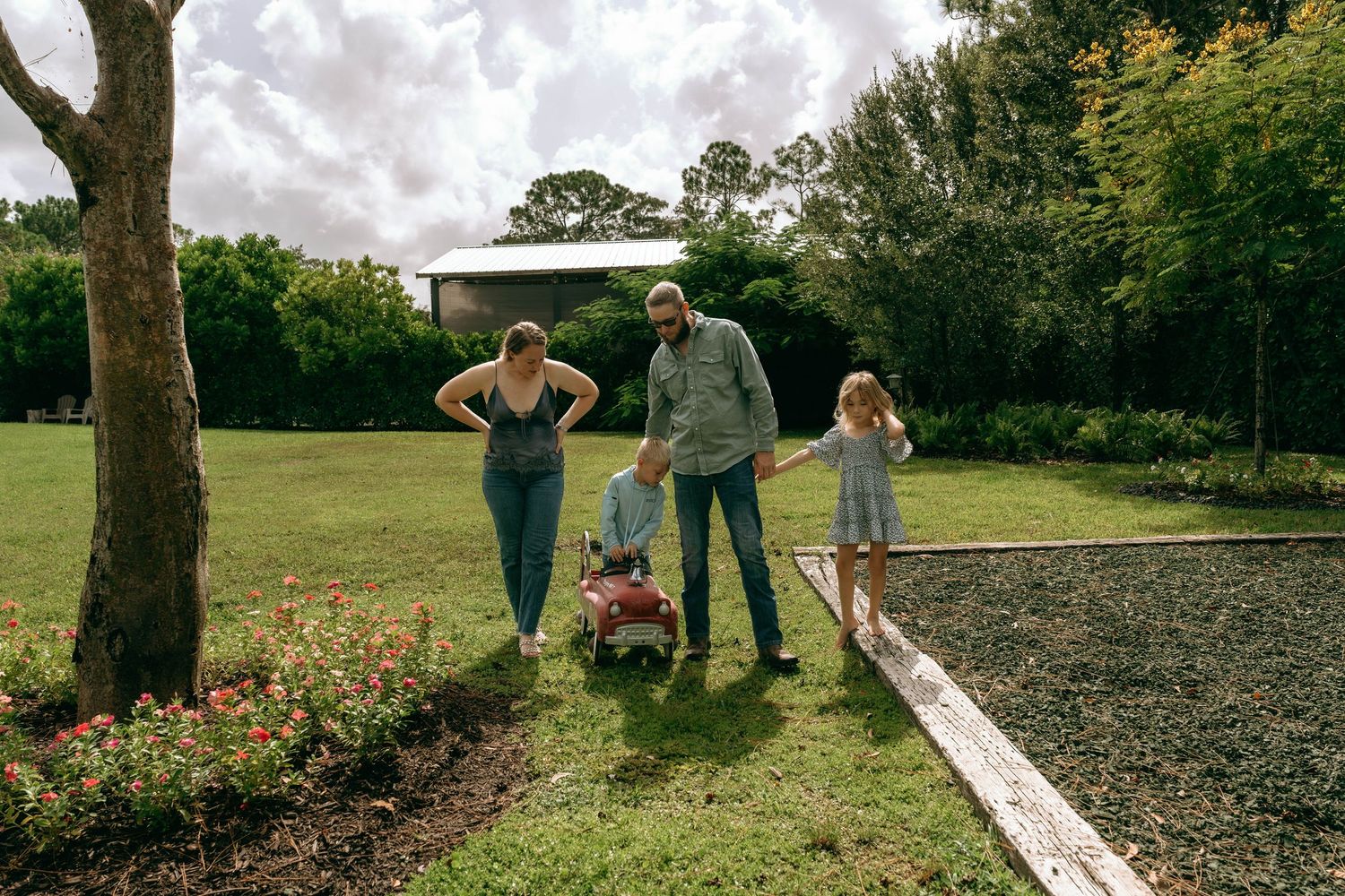 Family of four enjoying a sunny day in a lush green backyard garden with a young child in a red wagon.