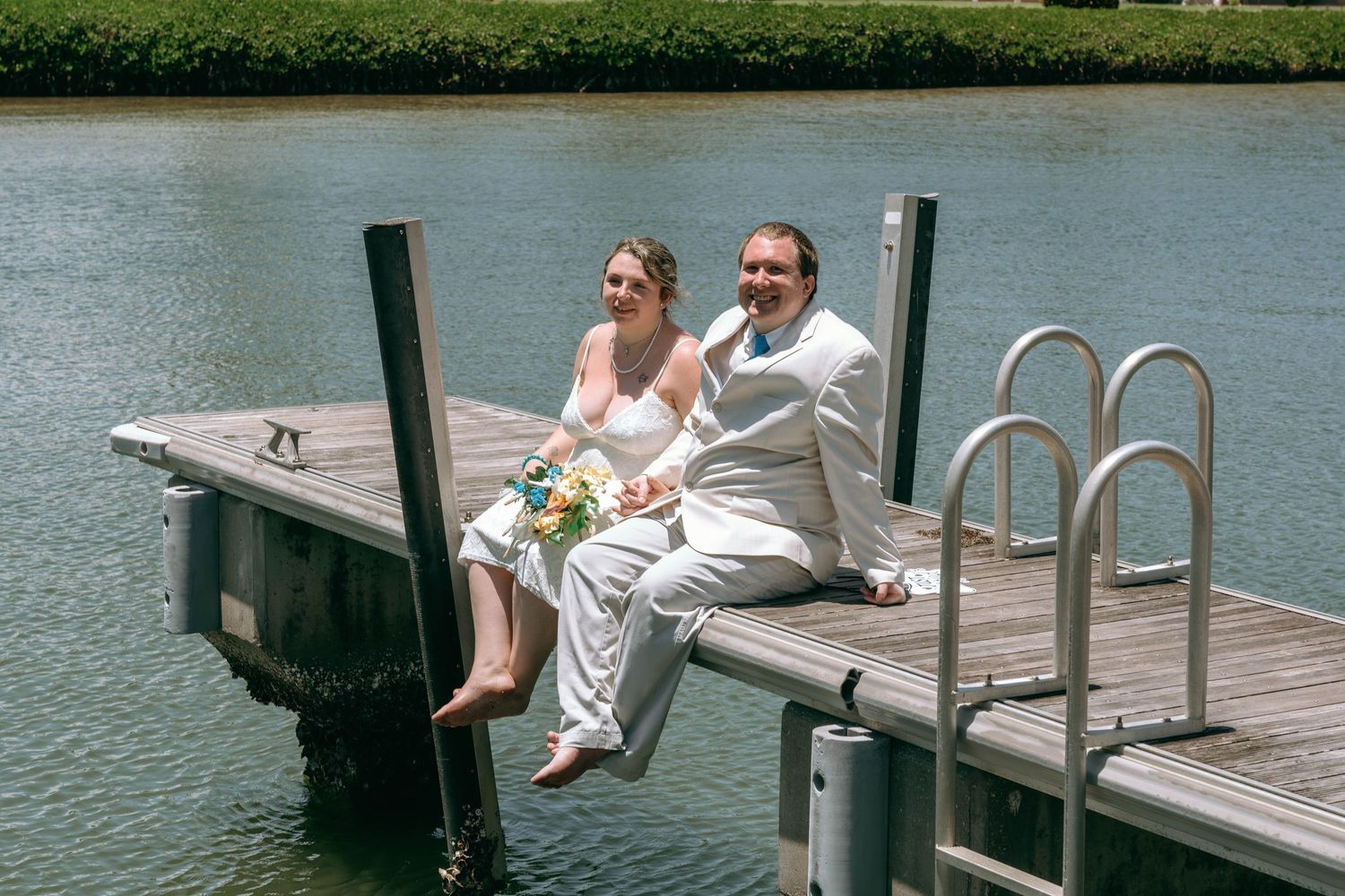 Bride and groom sitting on a wooden dock by the water, smiling and holding a bouquet on their wedding day.