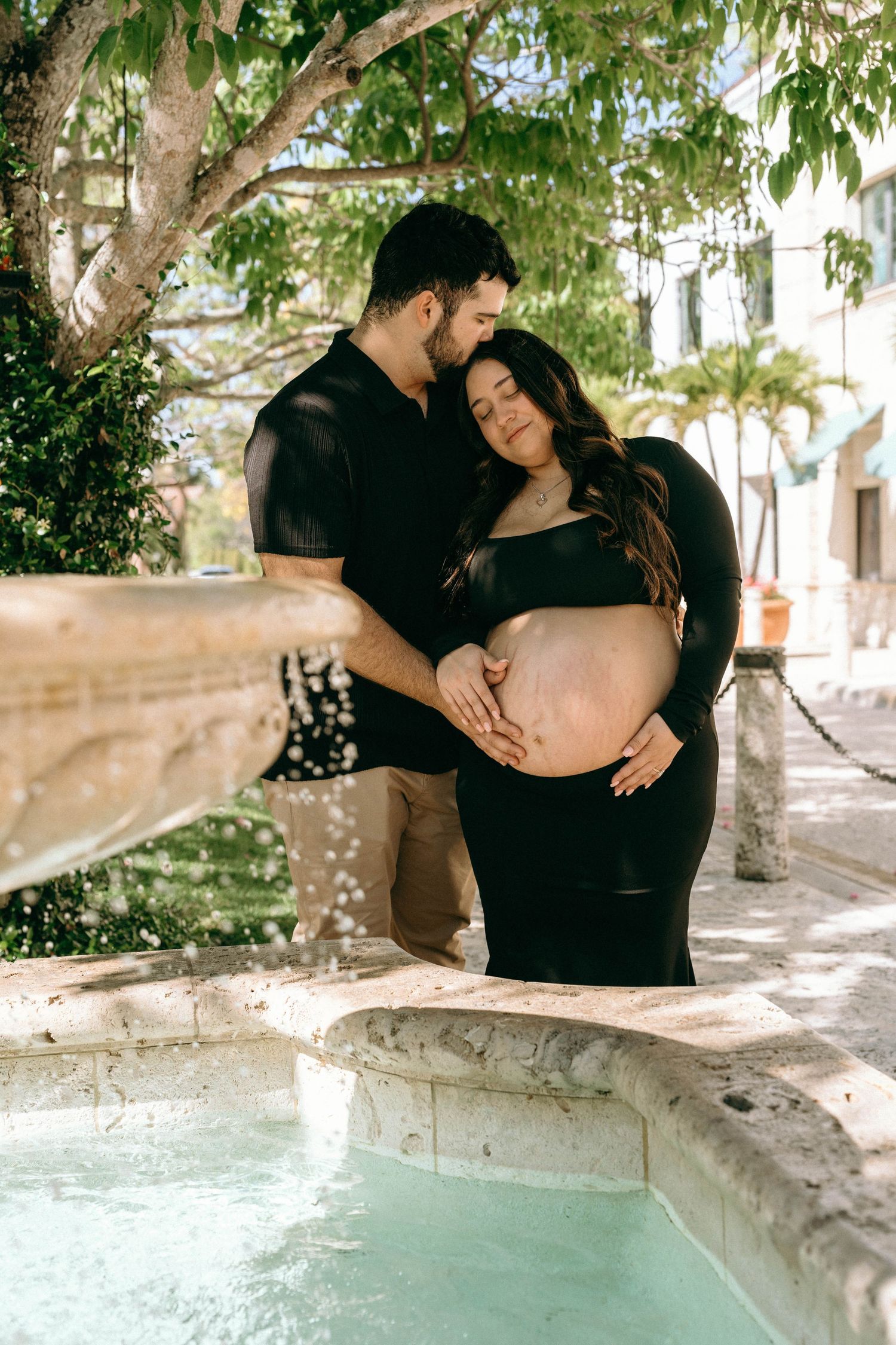 Couple tenderly cradling pregnant belly by a fountain outdoors, surrounded by lush greenery in a maternity photo shoot. Worth Avenue, Palm B