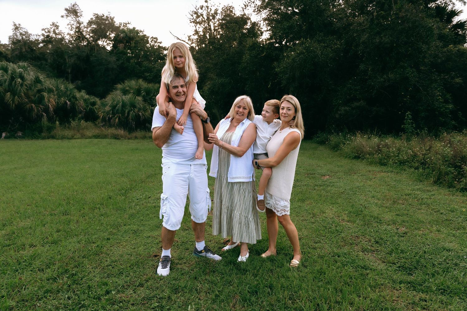 A happy family of five poses together on a green lawn surrounded by lush trees at golden hour Riverbend Park