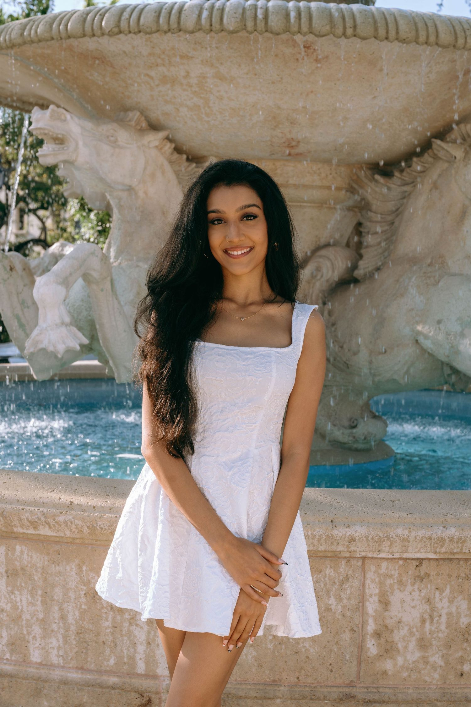 Young woman in white mini dress smiling by ornate stone fountain on Worth Avenue Palm beach.