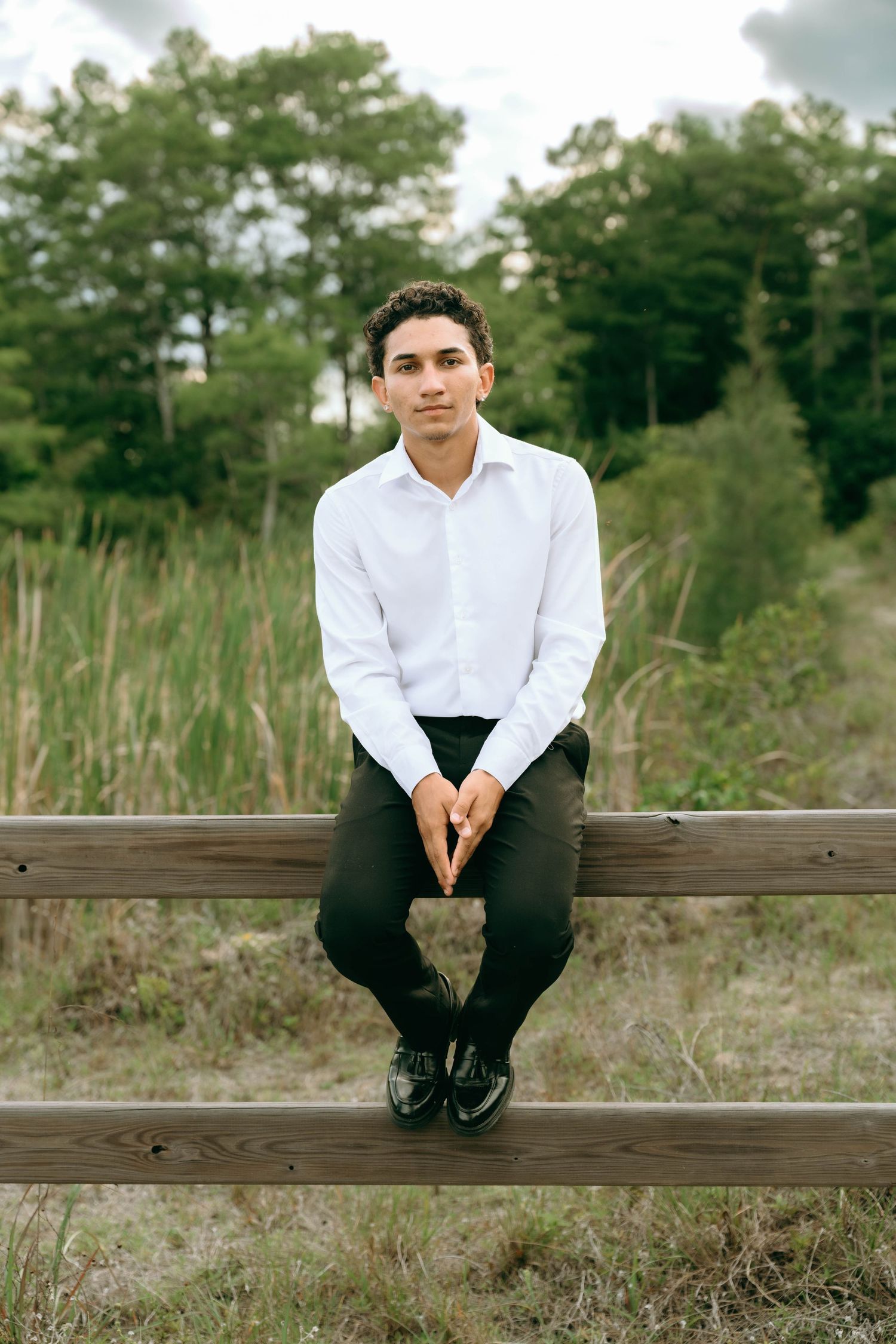 A young man in a white dress shirt and black pants sits on a wooden fence in a natural outdoor setting. Senior Photos at Colt Farms