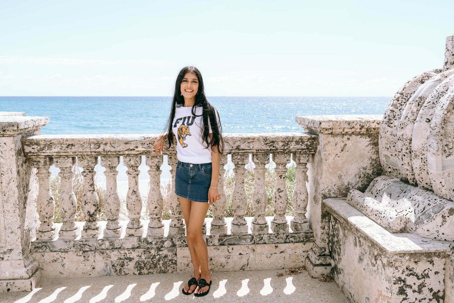 Young woman in FIU shirt and denim skirt poses on stone balcony overlooking blue ocean on Worth Avenue Palm beach.