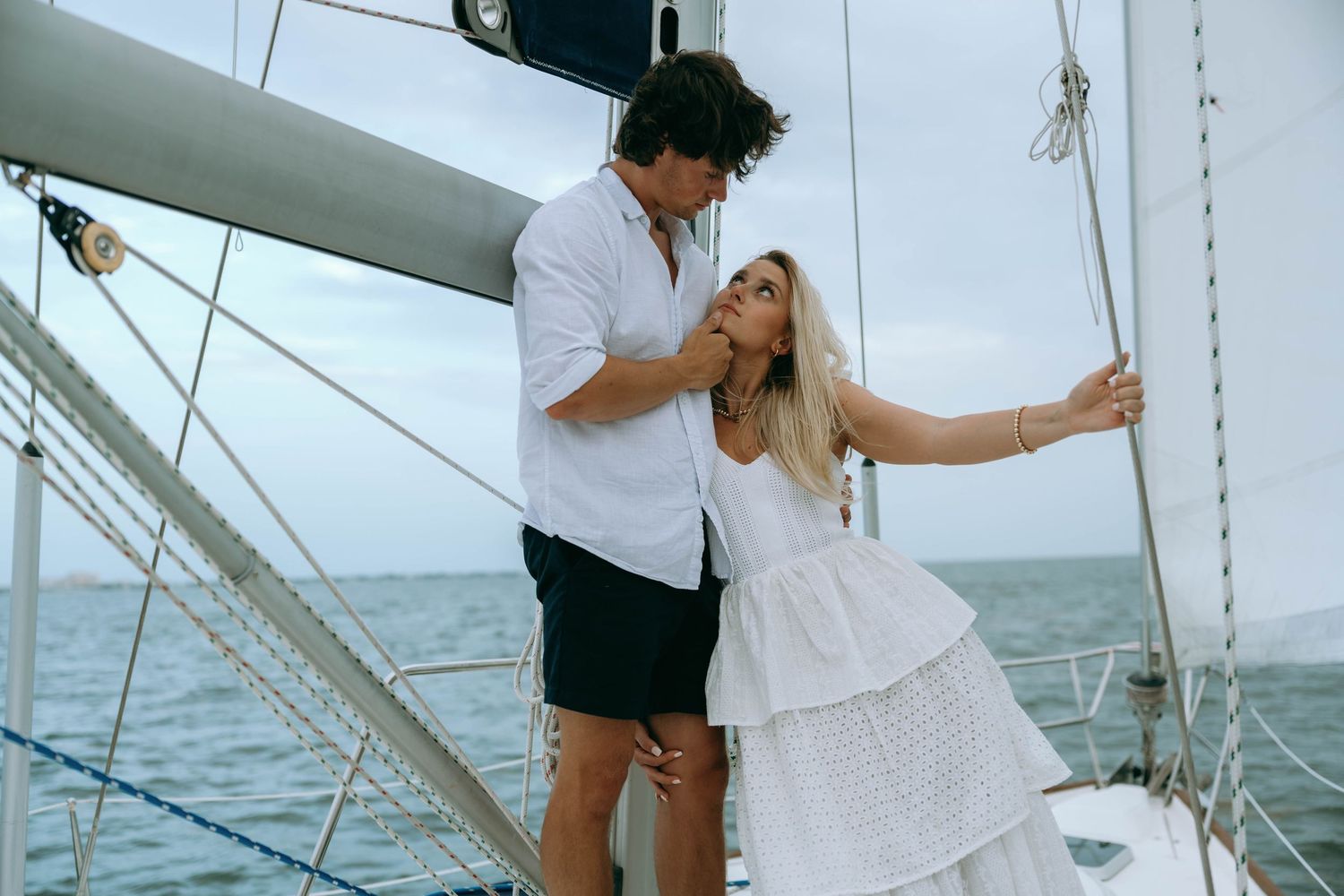 Couple sharing a romantic moment on a sailboat deck with ocean views, woman in white dress holding mast.