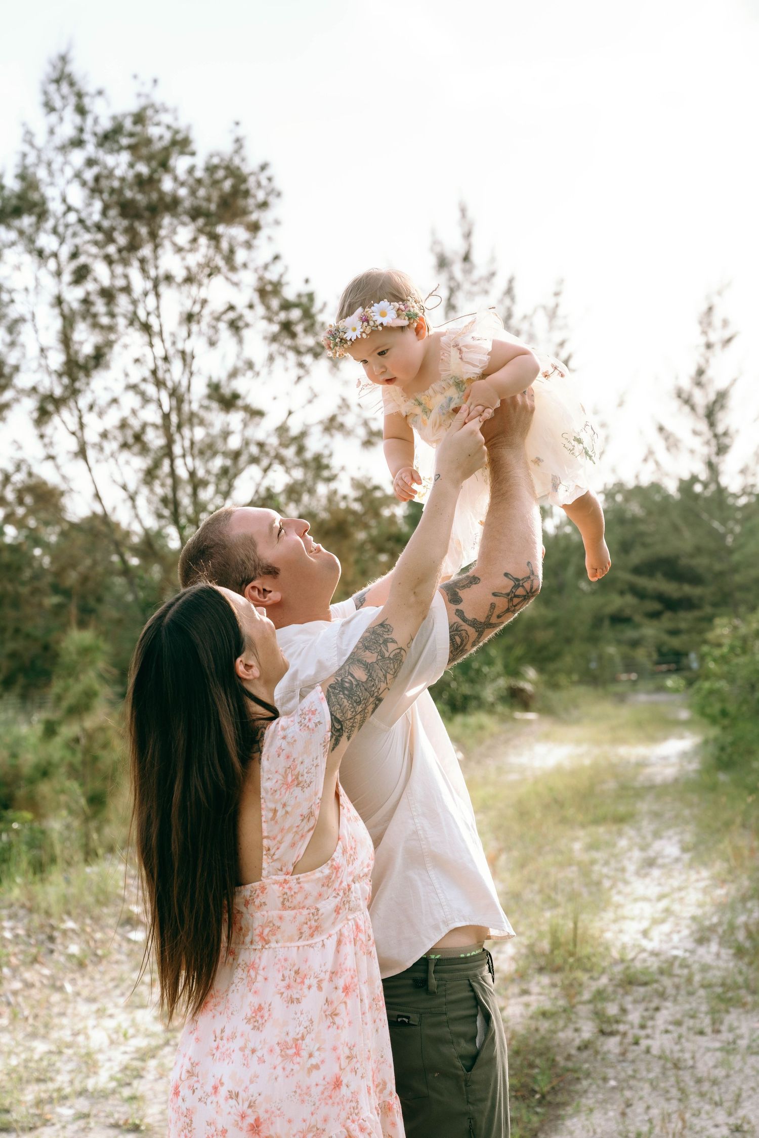 A tattooed father lifts his baby girl in a floral headband while mother watches, outdoors in a sunny forest path.