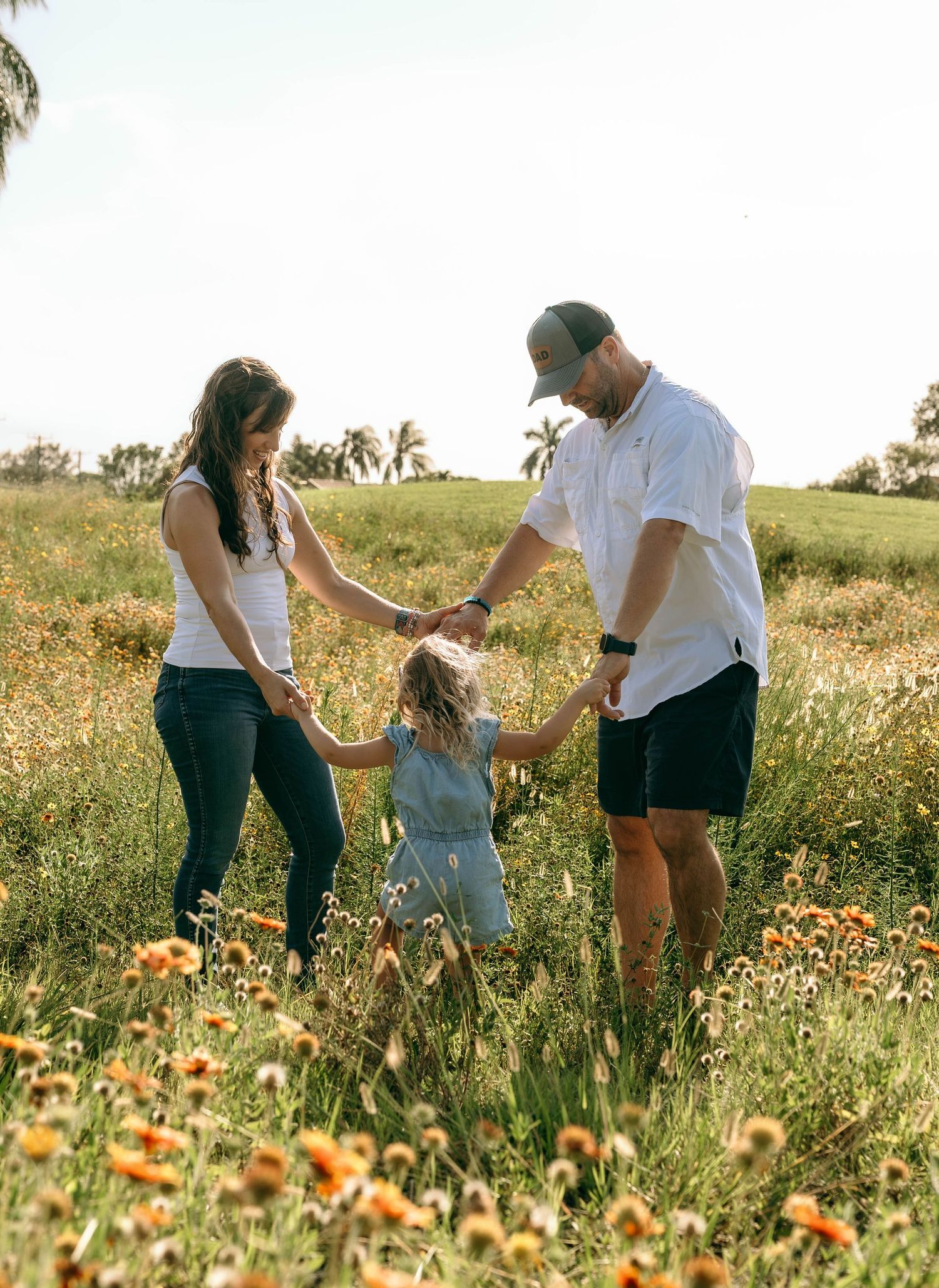 Family of three holding hands in a sunny wildflower meadow, parents swinging young child between them.