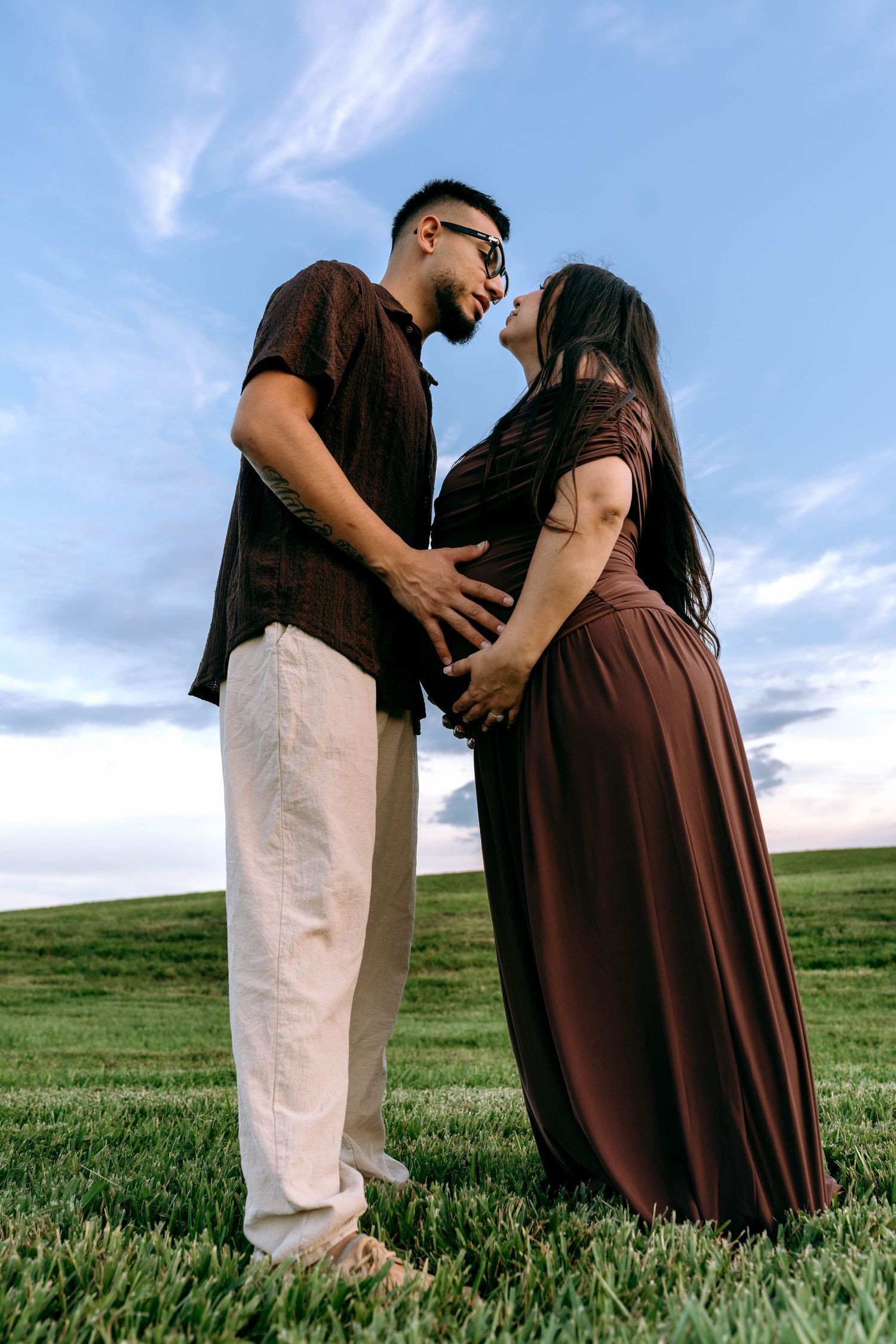 Couple in matching brown outfits sharing a romantic moment on a green field, woman in maxi dress with baby bump.
