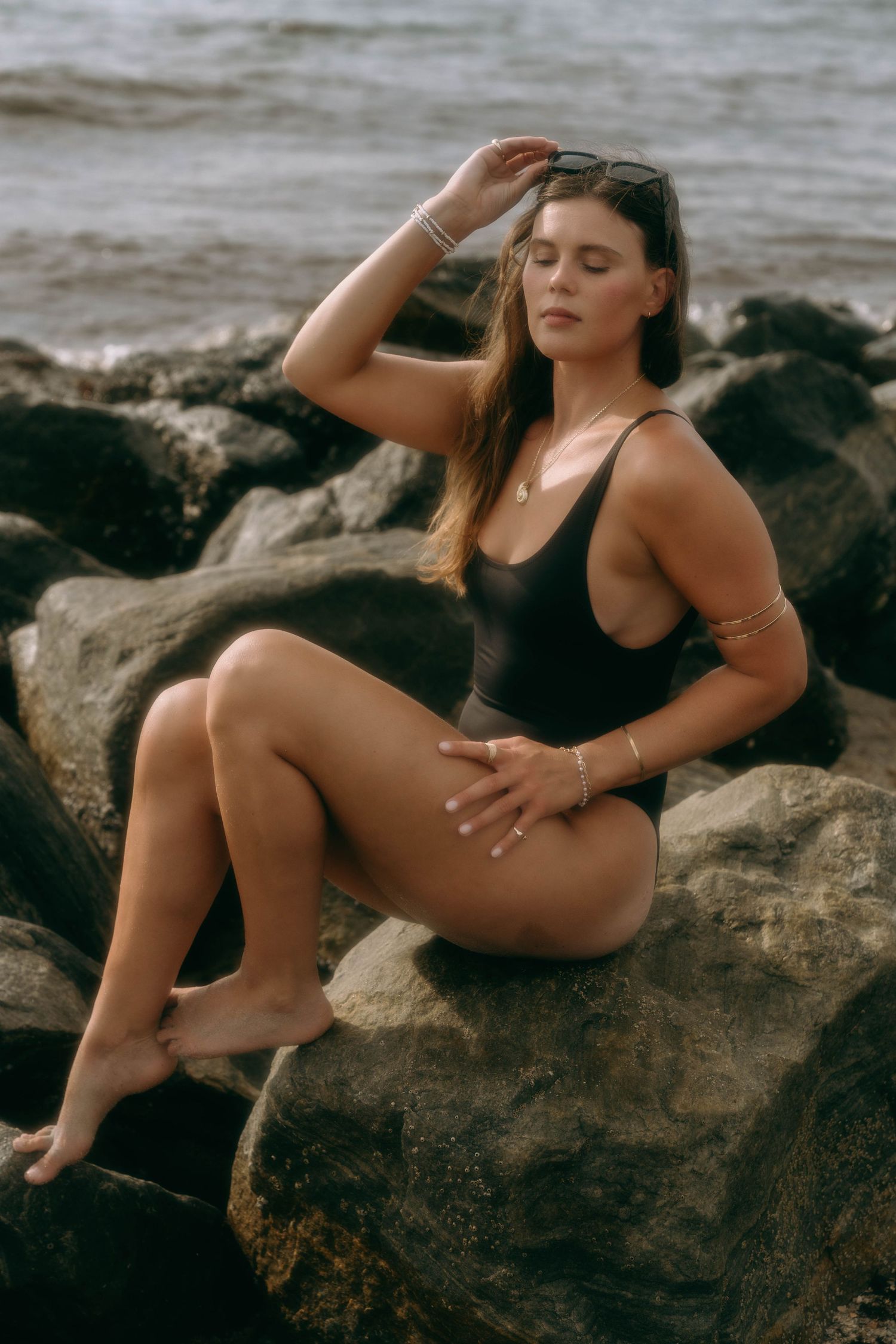 Woman in black one-piece swimsuit sitting on rocks by the ocean.