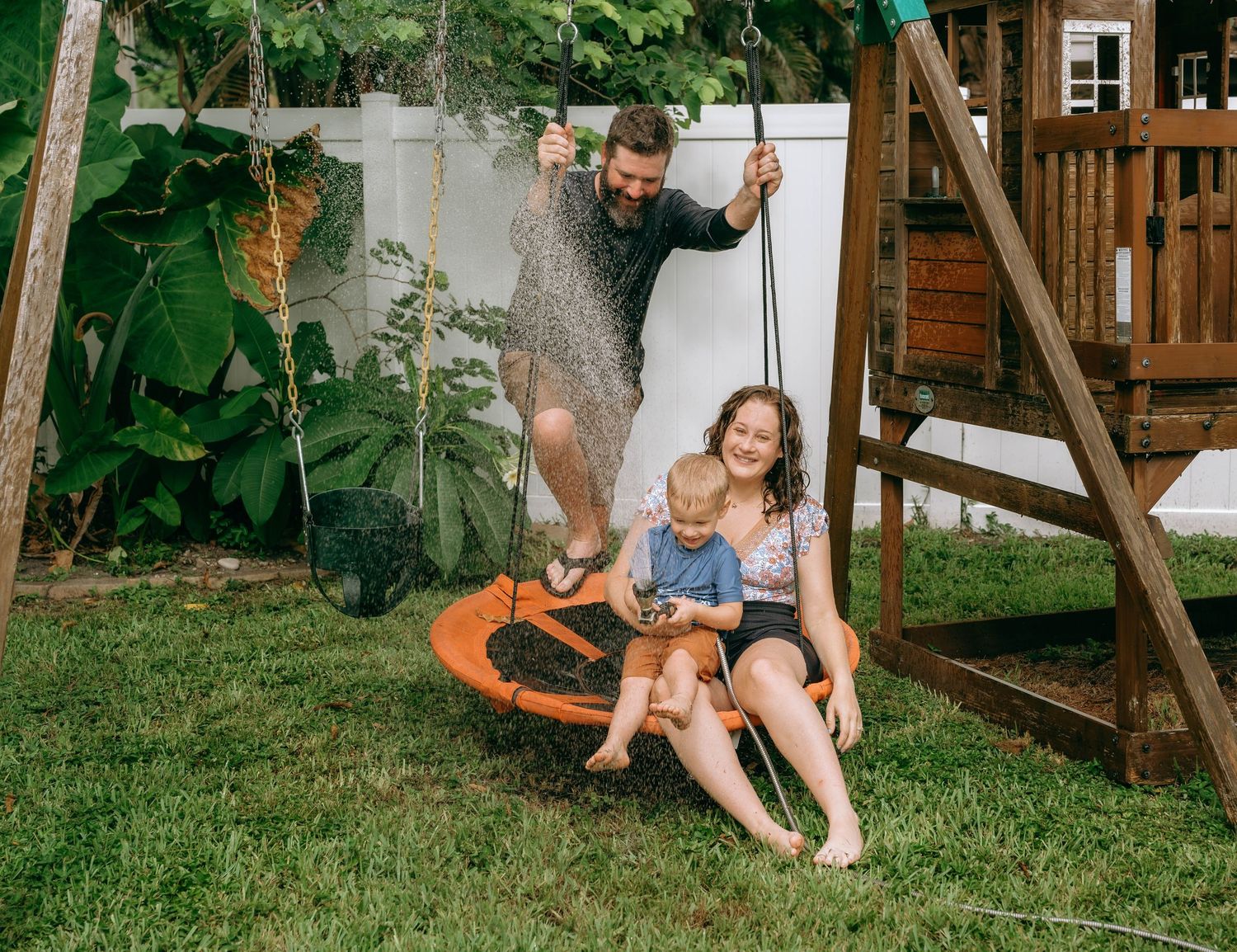 Family playing in backyard with man spraying water hose on woman and child sitting on orange swing set.