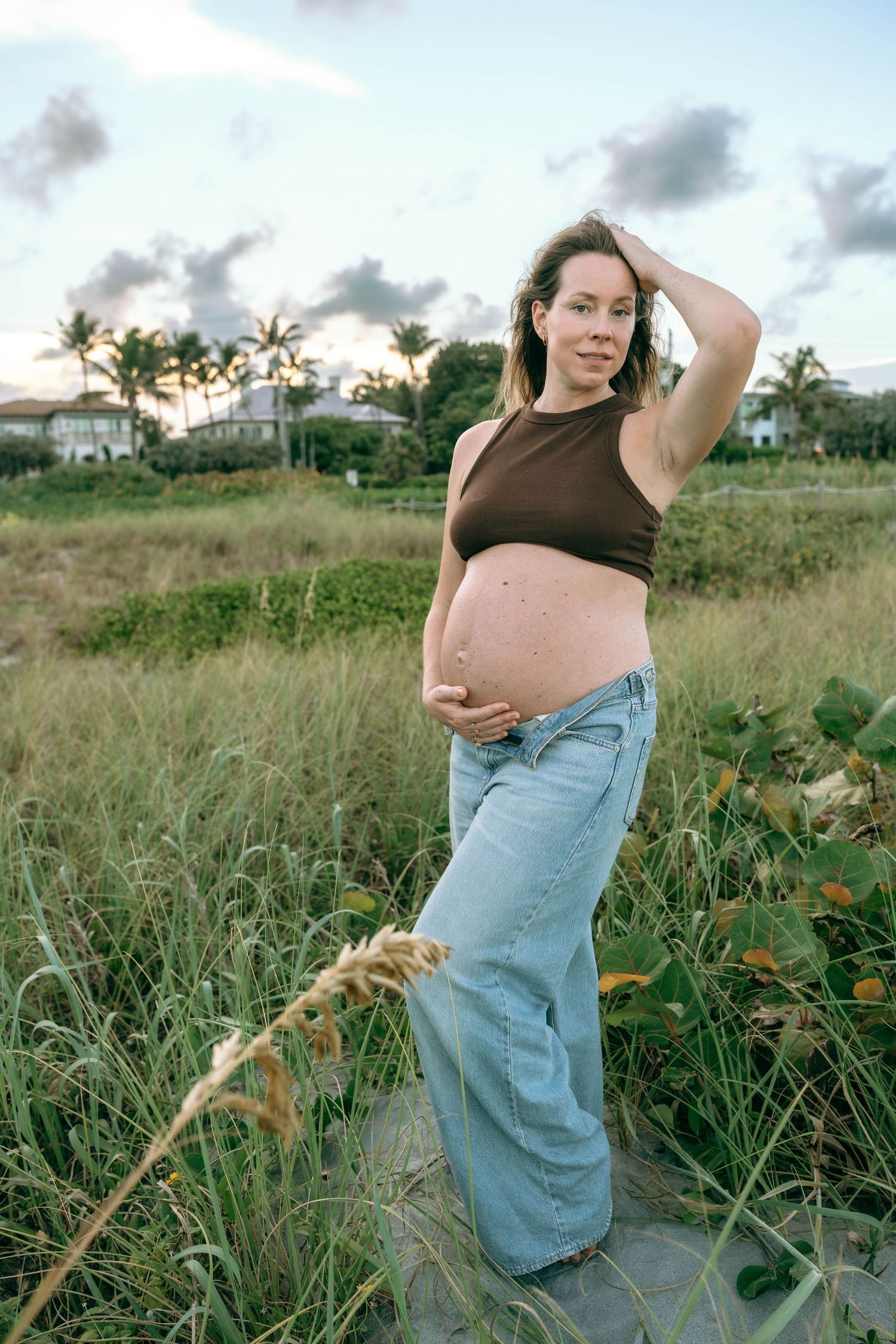 Pregnant woman in brown crop top and jeans poses in lush beach grass at dusk with palm trees behind her.