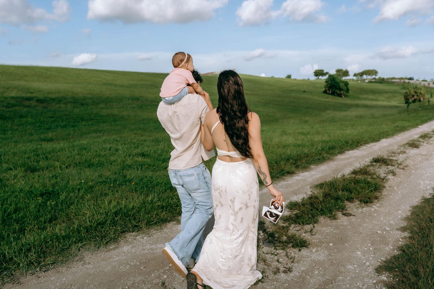 A couple walking along a countryside dirt path, man carrying a baby while woman holds a hat.