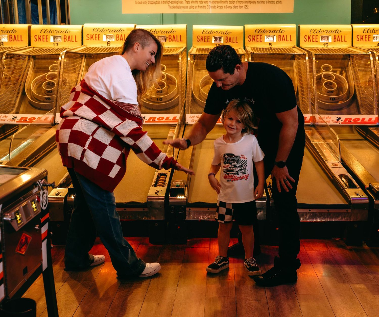 Two adults help a young child play skee-ball at an indoor arcade with wooden lanes.