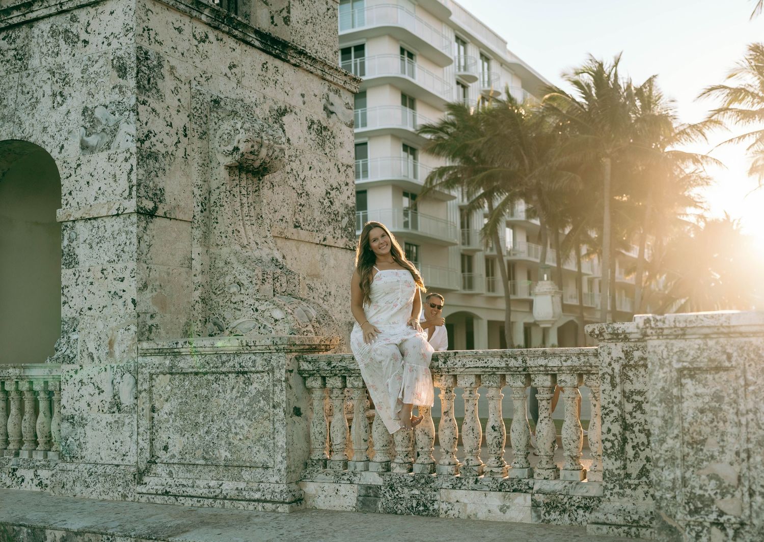 Woman in white dress poses on stone balustrade with palm trees and golden sunset in Miami.