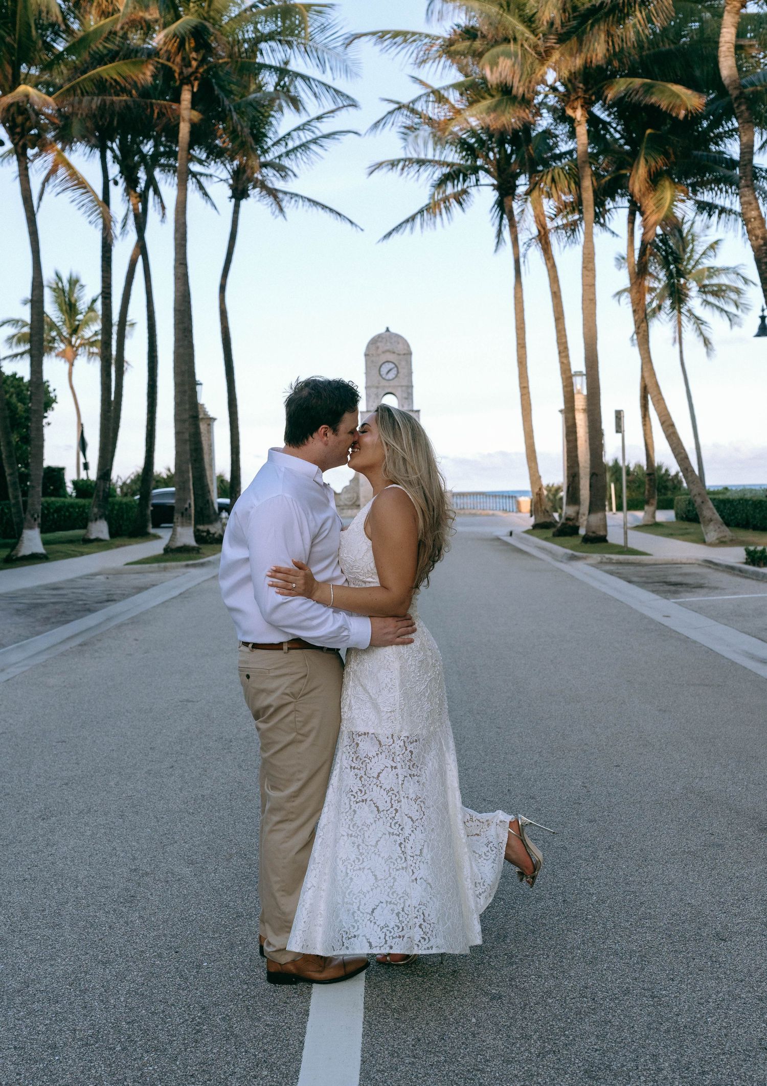 Couple sharing a kiss on a palm-lined road with a clock tower at sunset.