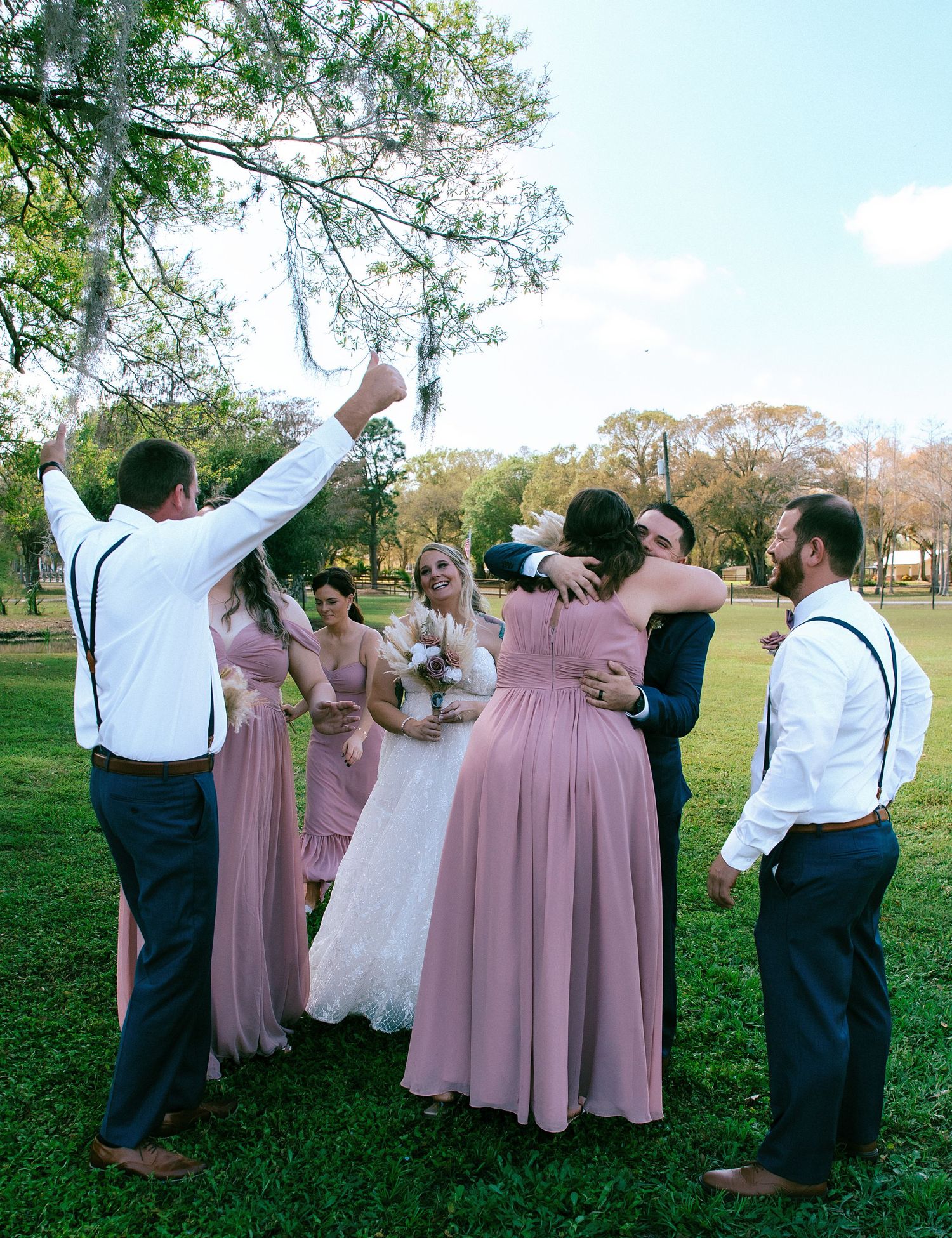 Wedding party celebrating outdoors in pink bridesmaid dresses and suspenders on lush green lawn.