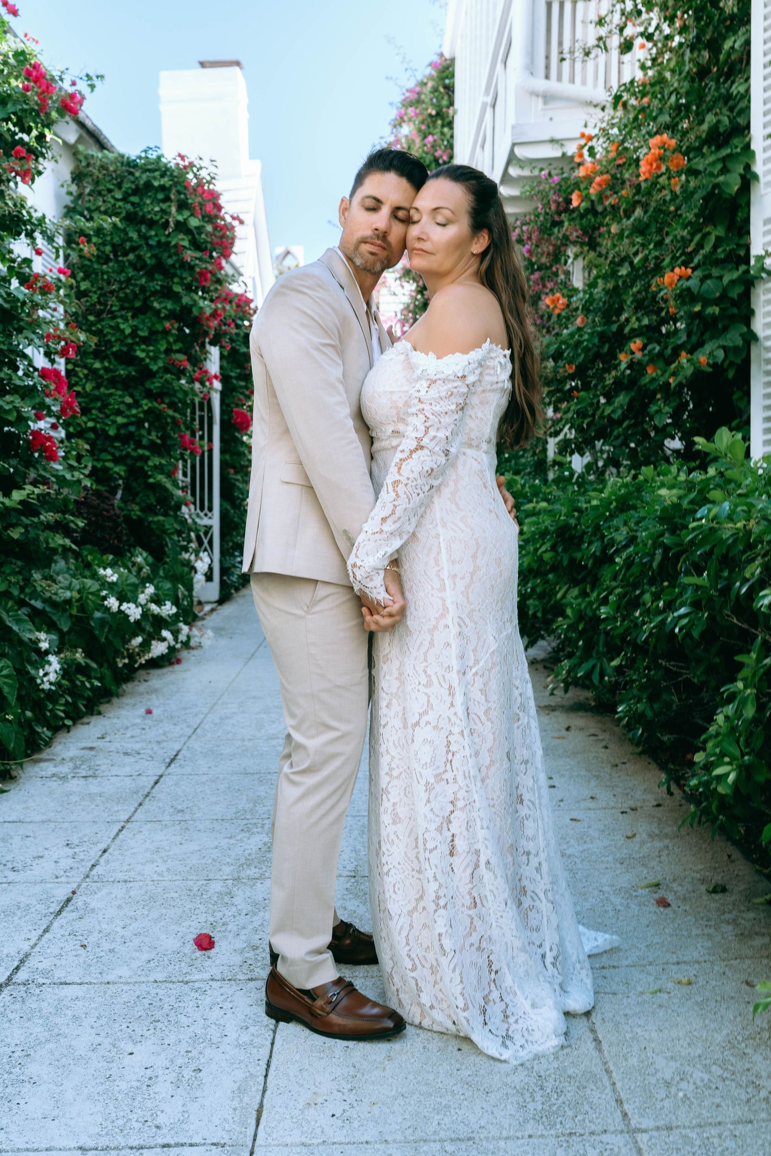 Couple in wedding attire share a romantic moment in a lush floral alleyway, bride in elegant lace gown.