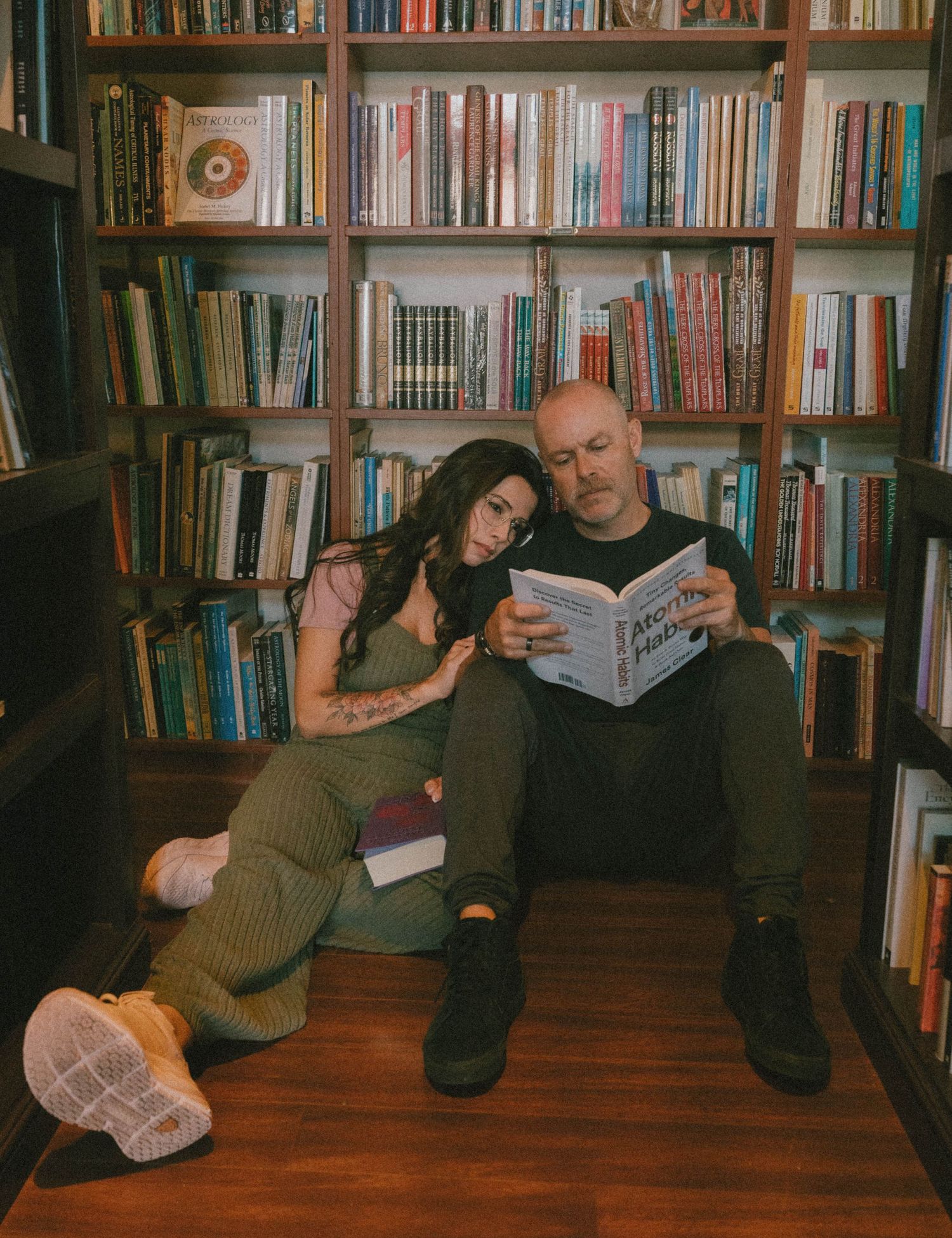 Couple sitting on floor reading book together in cozy library with floor-to-ceiling bookshelves.
