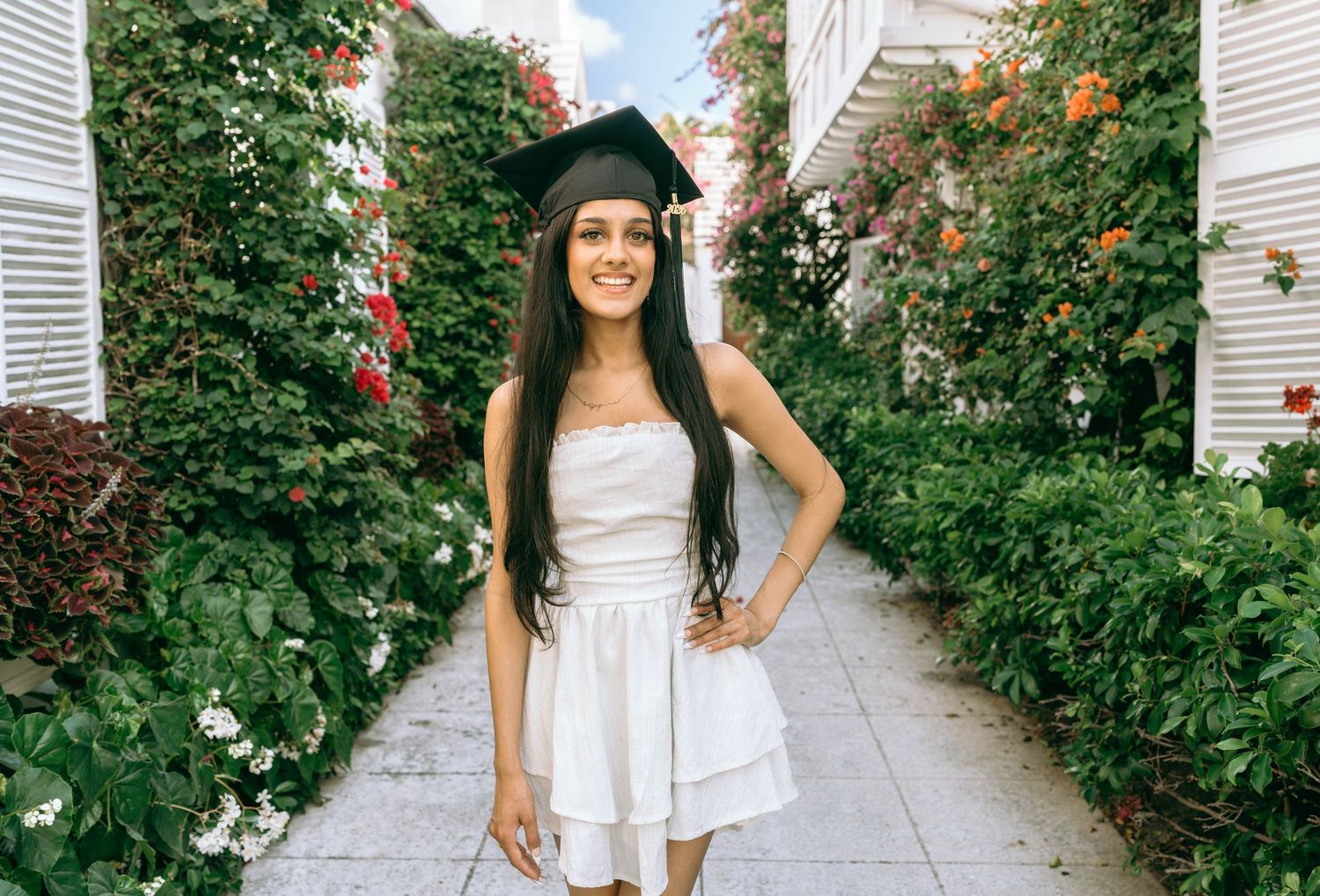 Young woman in white dress and graduation cap smiles outdoors among lush green hedges and colorful flowers on Worth Avenue Palm beach.