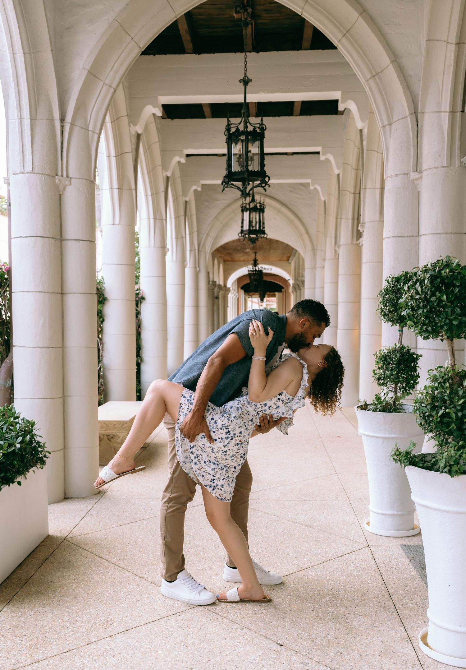 Romantic couple sharing a dip kiss in an elegant arched colonnade with white columns and topiary plants. Engagement photos on Worth Avenue