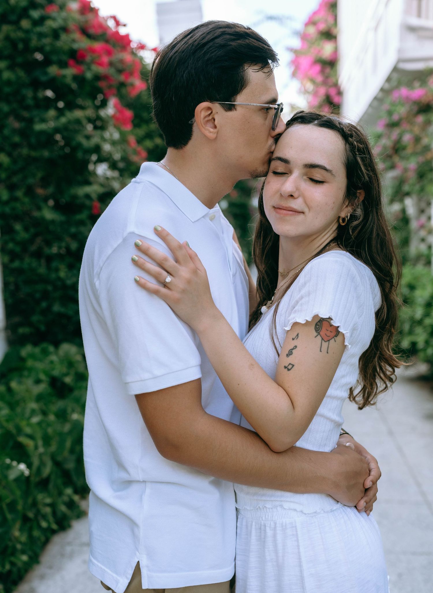 A couple in white outfits embracing outdoors, man kissing woman's forehead near colorful flowers. Engagement photos on Worth Avenue