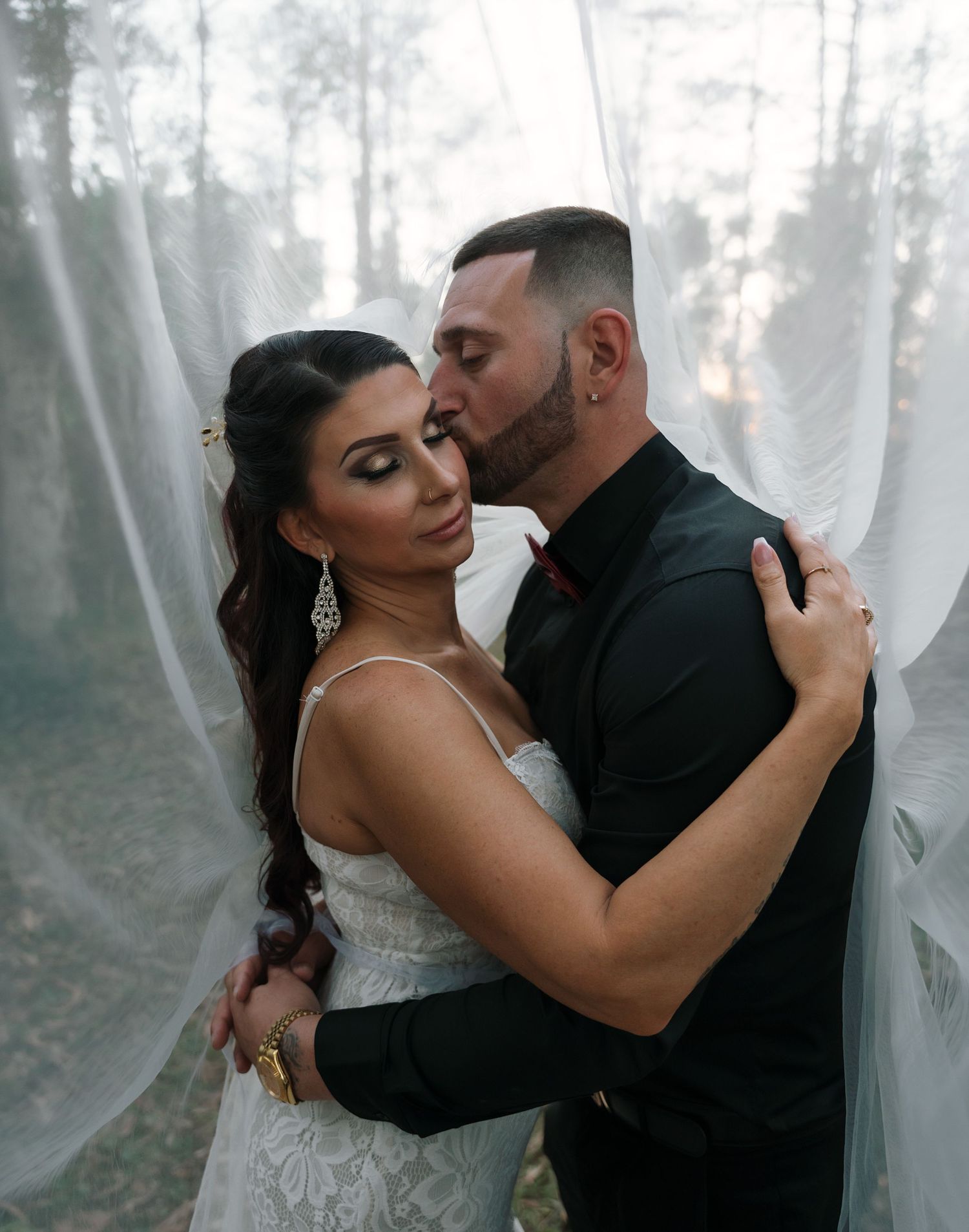 A groom in black kisses his bride in a white lace gown, embracing under flowing white fabric in a misty forest. Wedding at Colt Farms