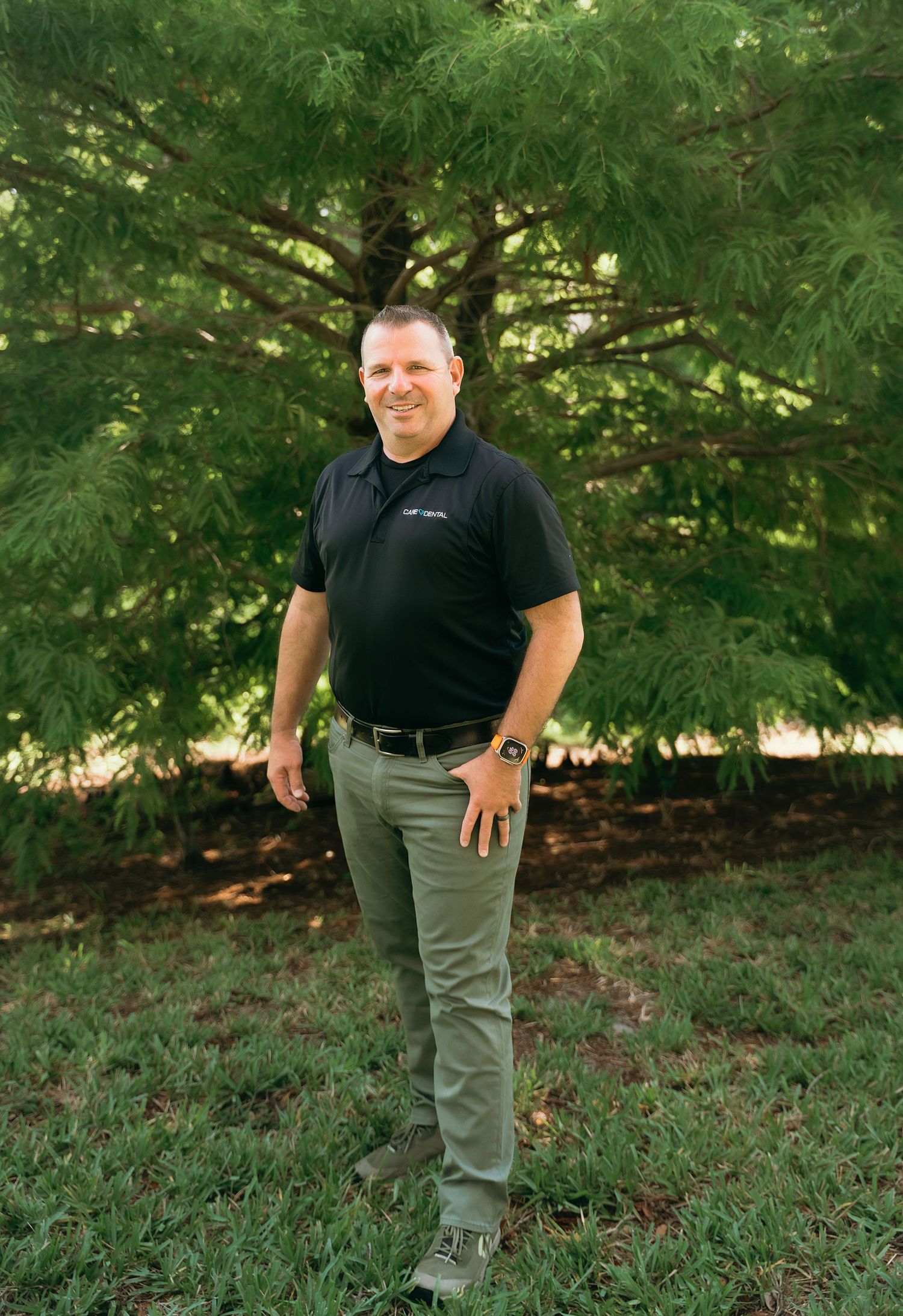 Full-length outdoor business portraits in black polo and khaki pants against trees.