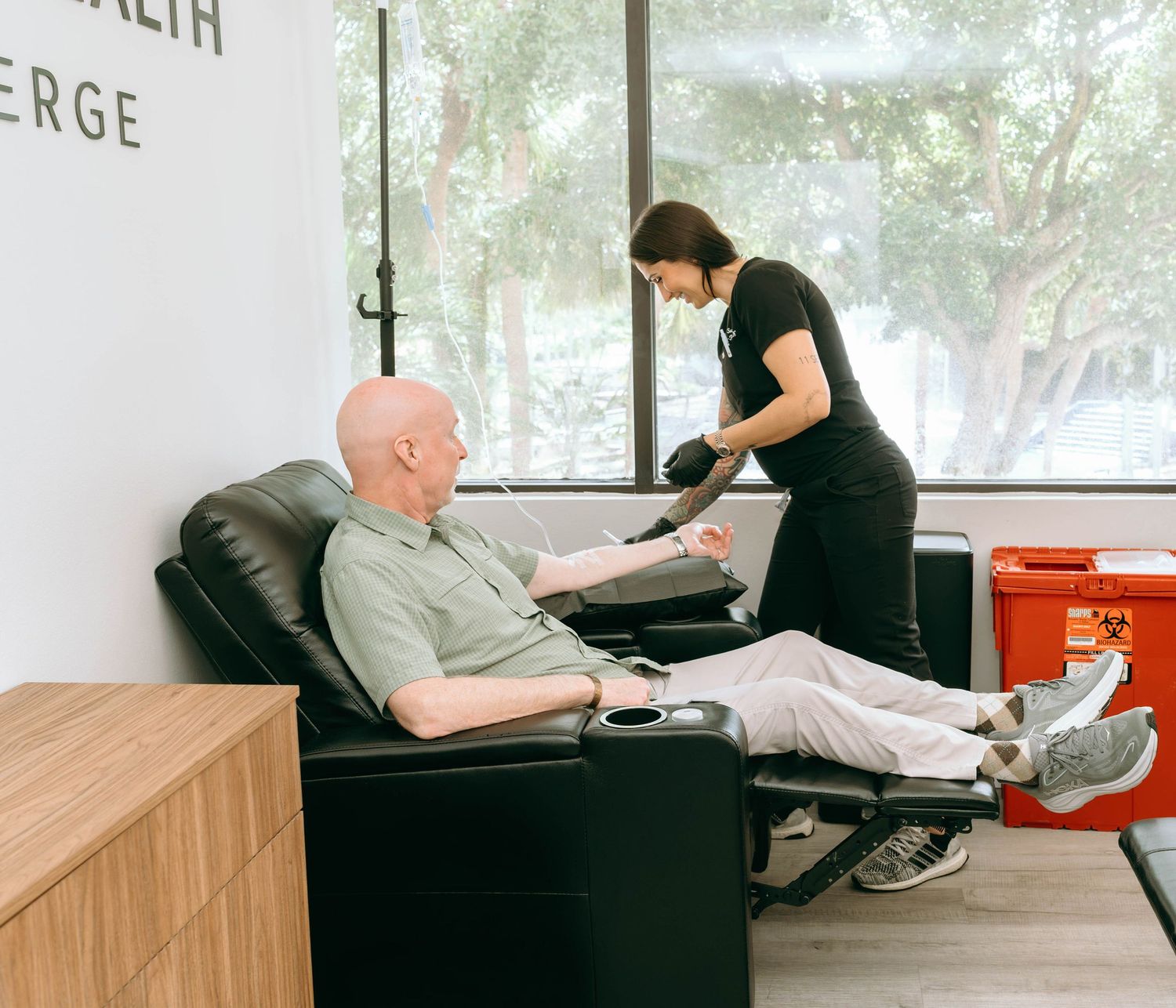 Healthcare worker drawing blood from a bald male patient seated in a black recliner chair at a medical concierge clinic.
